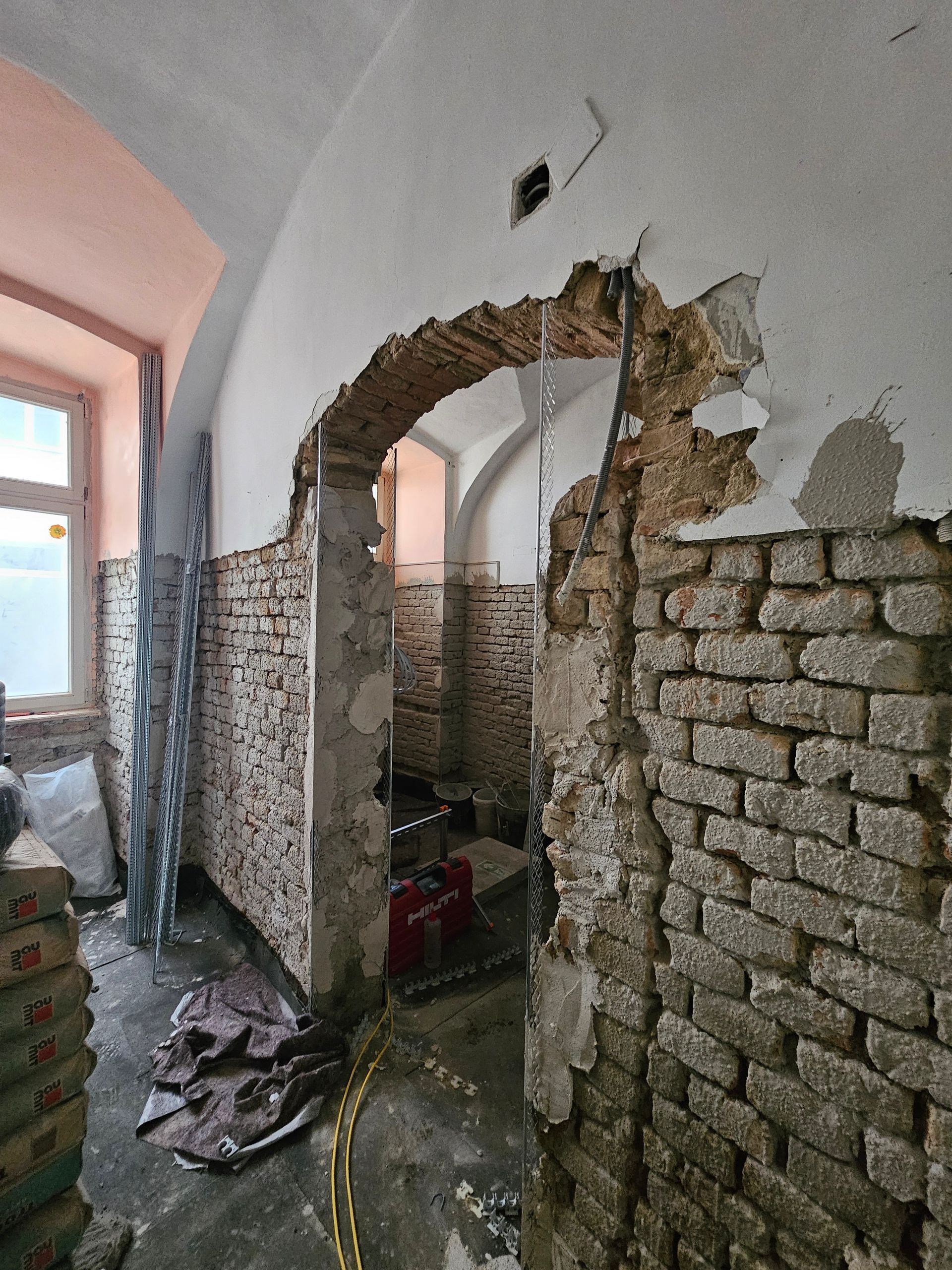 Ein gewölbter Türbogen mit freiliegenden Ziegeln und abblätterndem Putz in einer Baustelle im Innenbereich in der Nähe eines Fensters.