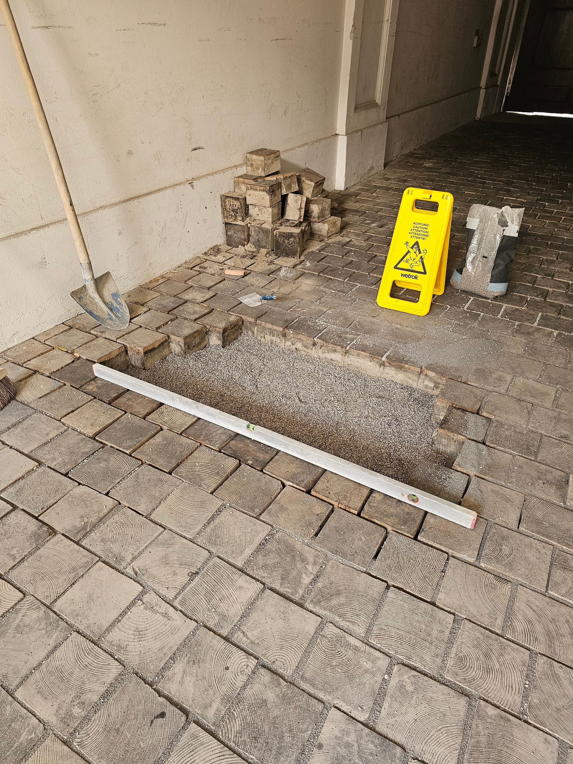 Ein offener Graben in einem Kopfsteinpflasterweg, über den eine Richtlatte verläuft, und ein gelbes Warnschild in der Nähe.