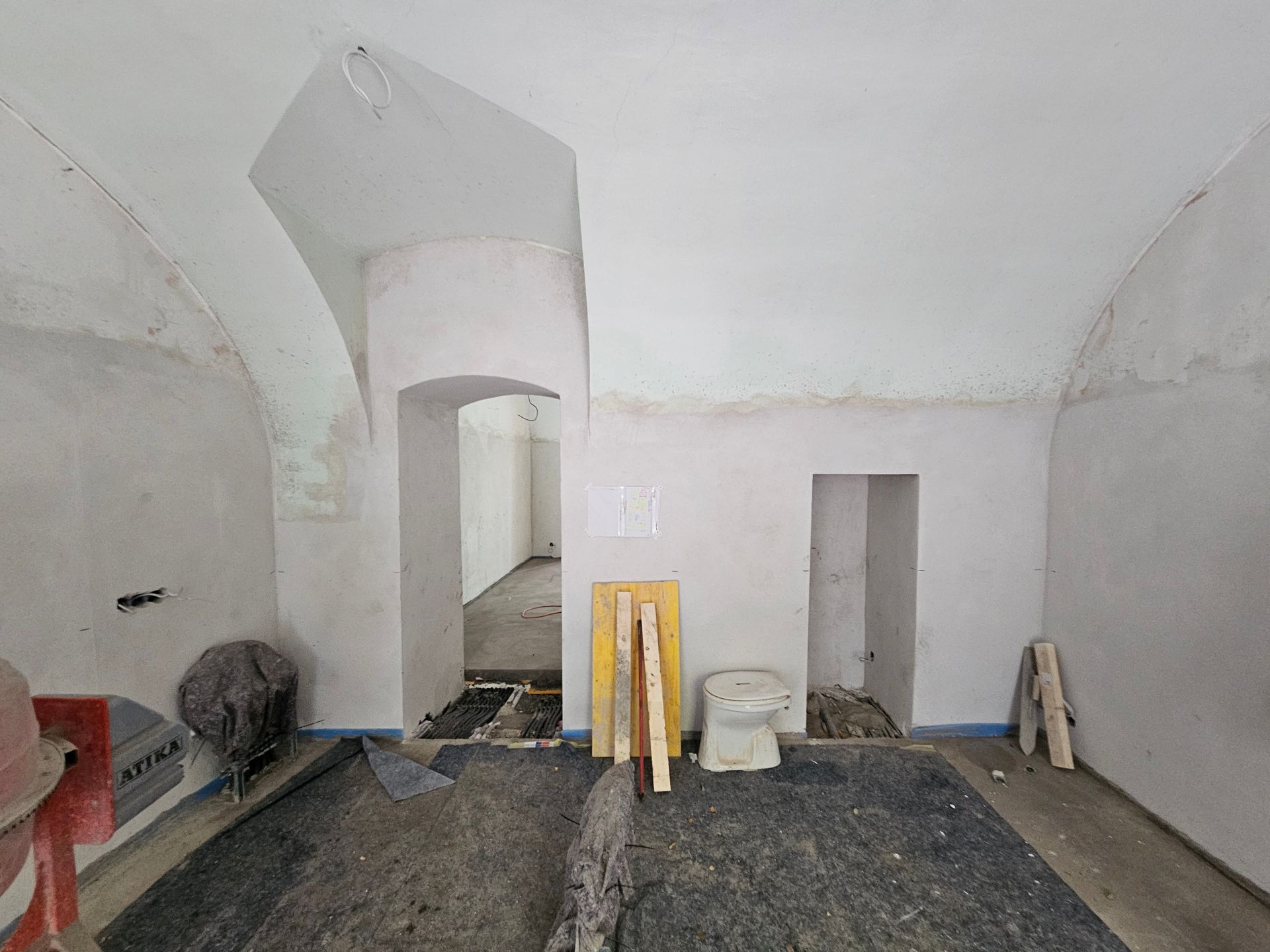 Ein unfertiger Kellerraum mit weißen, gewölbten Wänden, zwei Türen, einer Toilette und Baumaterialien auf dem Boden.