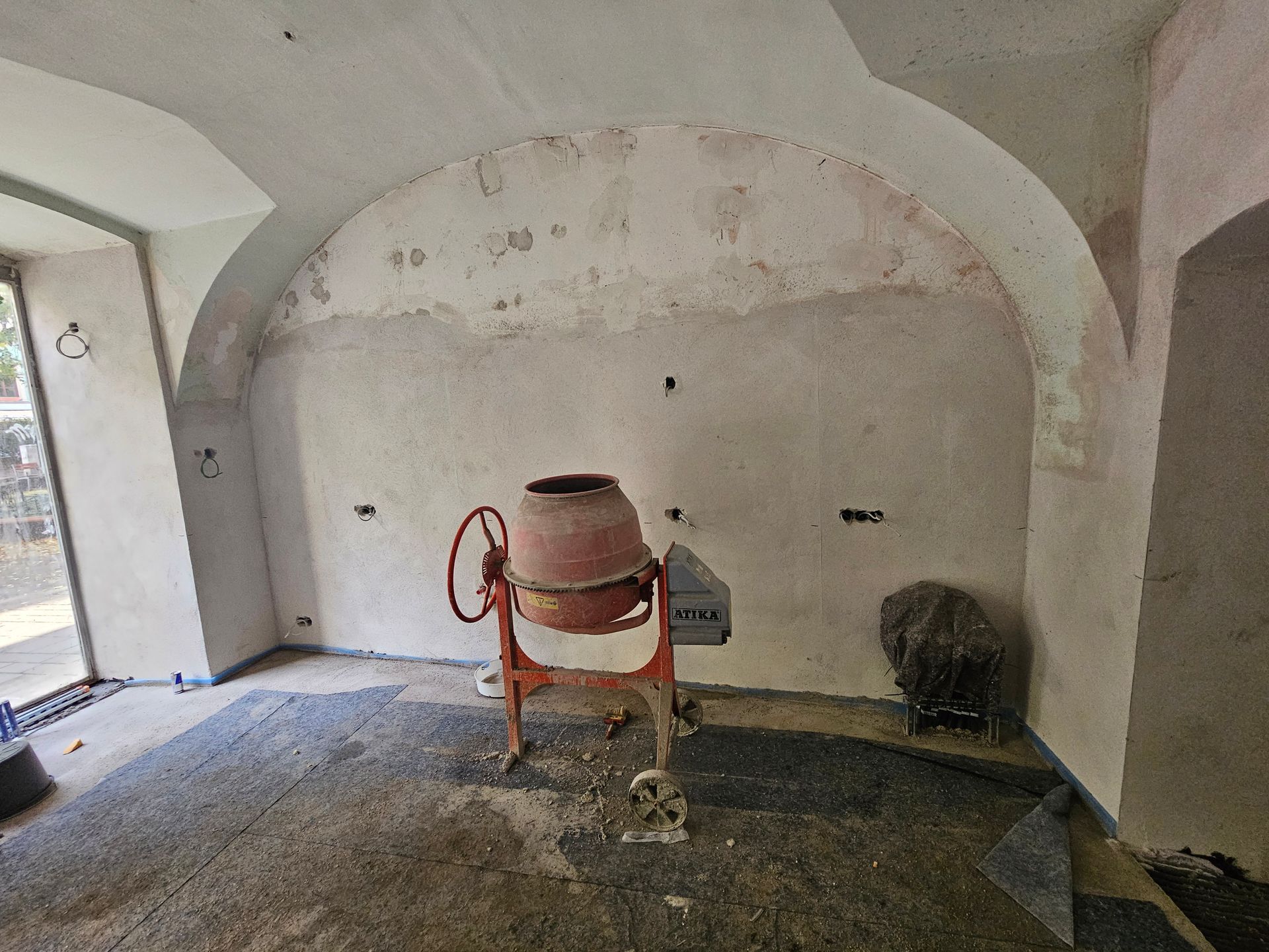In einem Raum mit gewölbten Decken, kahlen Wänden und einem mit Bauschutt bedeckten Boden steht ein roter Betonmischer.