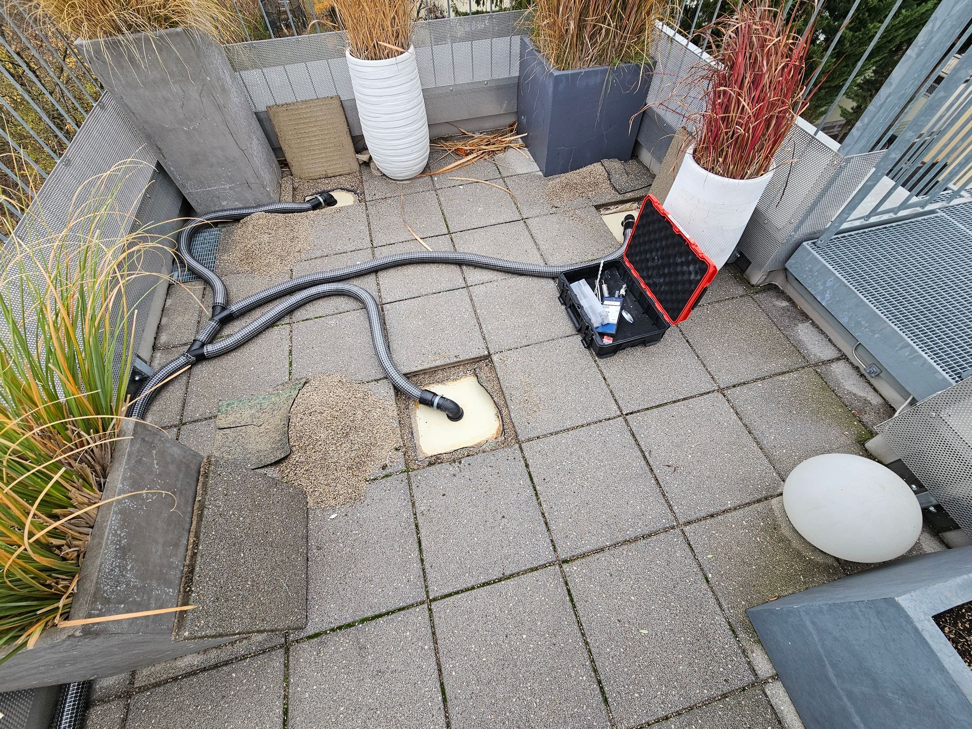 Eine Terrasse mit grauen Terrassenfliesen, mehreren Topfpflanzen und einem industriell anmutenden Trocknungsgerät mit an eine Entlüftung angeschlossenen Schläuchen.