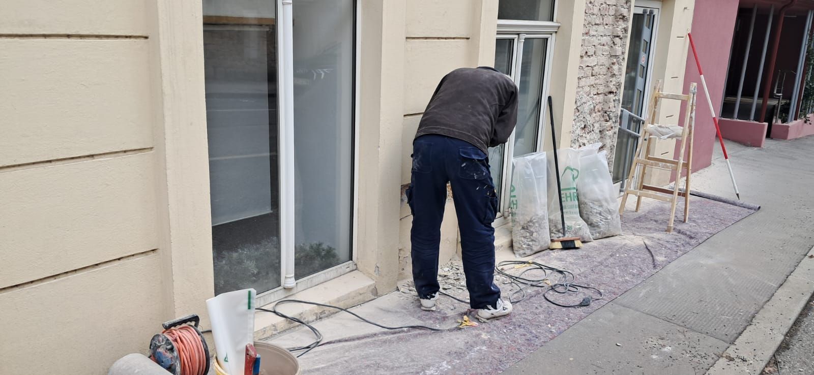 Eine Person in dunkler Arbeitskleidung steht vor einem Gebäude und führt mit einem Elektrowerkzeug Bau- oder Reparaturarbeiten durch.