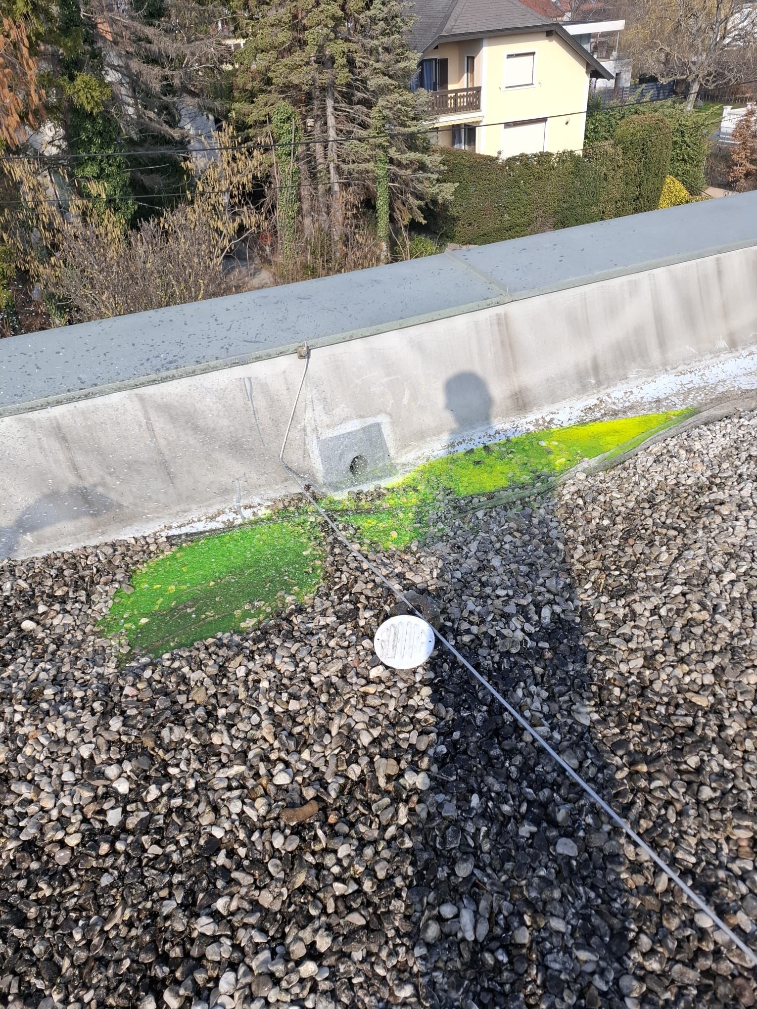 Eine leuchtend grüne Flüssigkeit ist ausgelaufen und hat den Kies auf einem Flachdach in der Nähe einer Betonmauer verfärbt; daneben befindet sich eine kleine weiße runde Markierung.