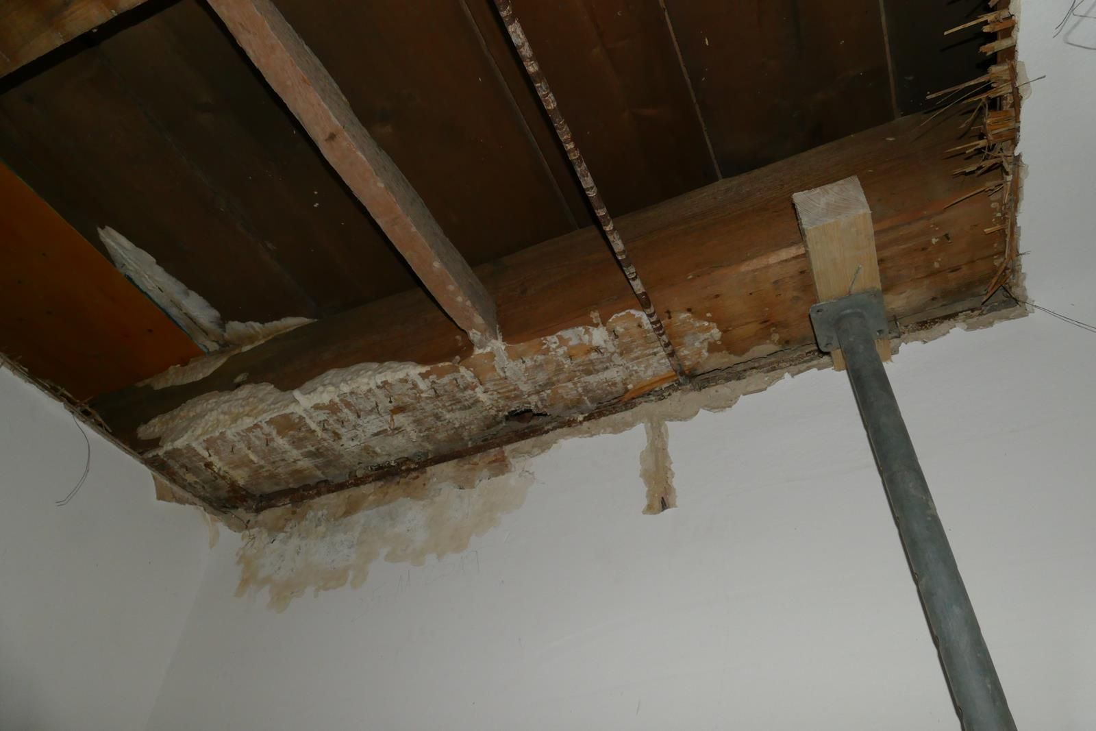 Ein tragender Metallpfosten stützt einen Holzbalken in einer Deckenöffnung, die sichtbare Wasserschäden und bröckelnden Putz aufweist.