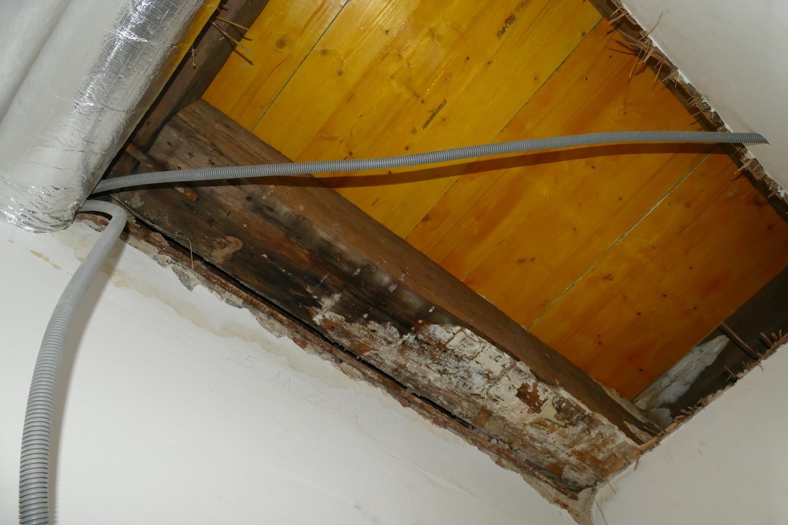 Ein freigelegter Teil der Decke gibt den Blick frei auf Holzplanken, dunkle, beschädigte Tragbalken und herabhängende elektrische Leitungen.