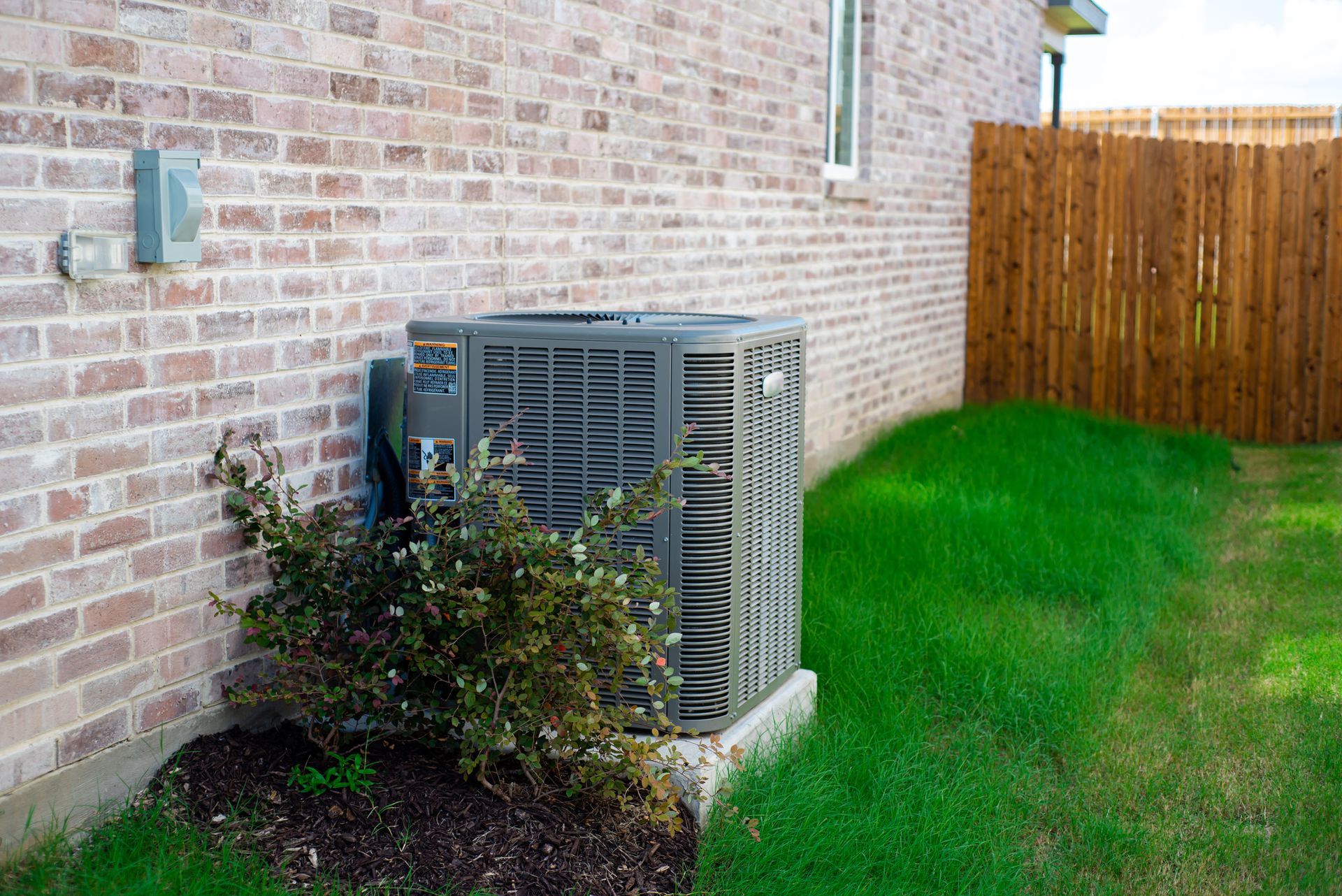 Outdoor AC unit beside brick house, partly hidden by shrubs, with green lawn and wooden fence nearby