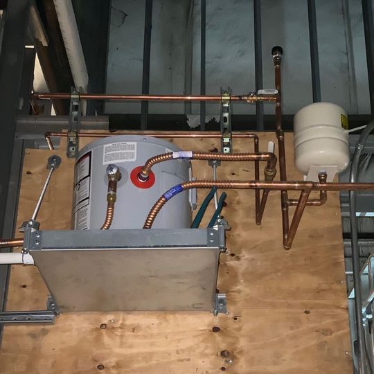 Industrial utility equipment with copper pipes, hoses, and a metal tank mounted against plywood and wall framing
