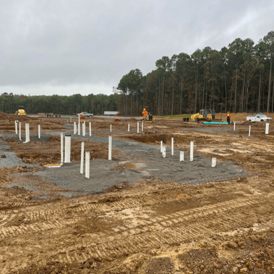 Construction site with concrete foundation slabs, white pipes, heavy equipment, and a tree line in the background