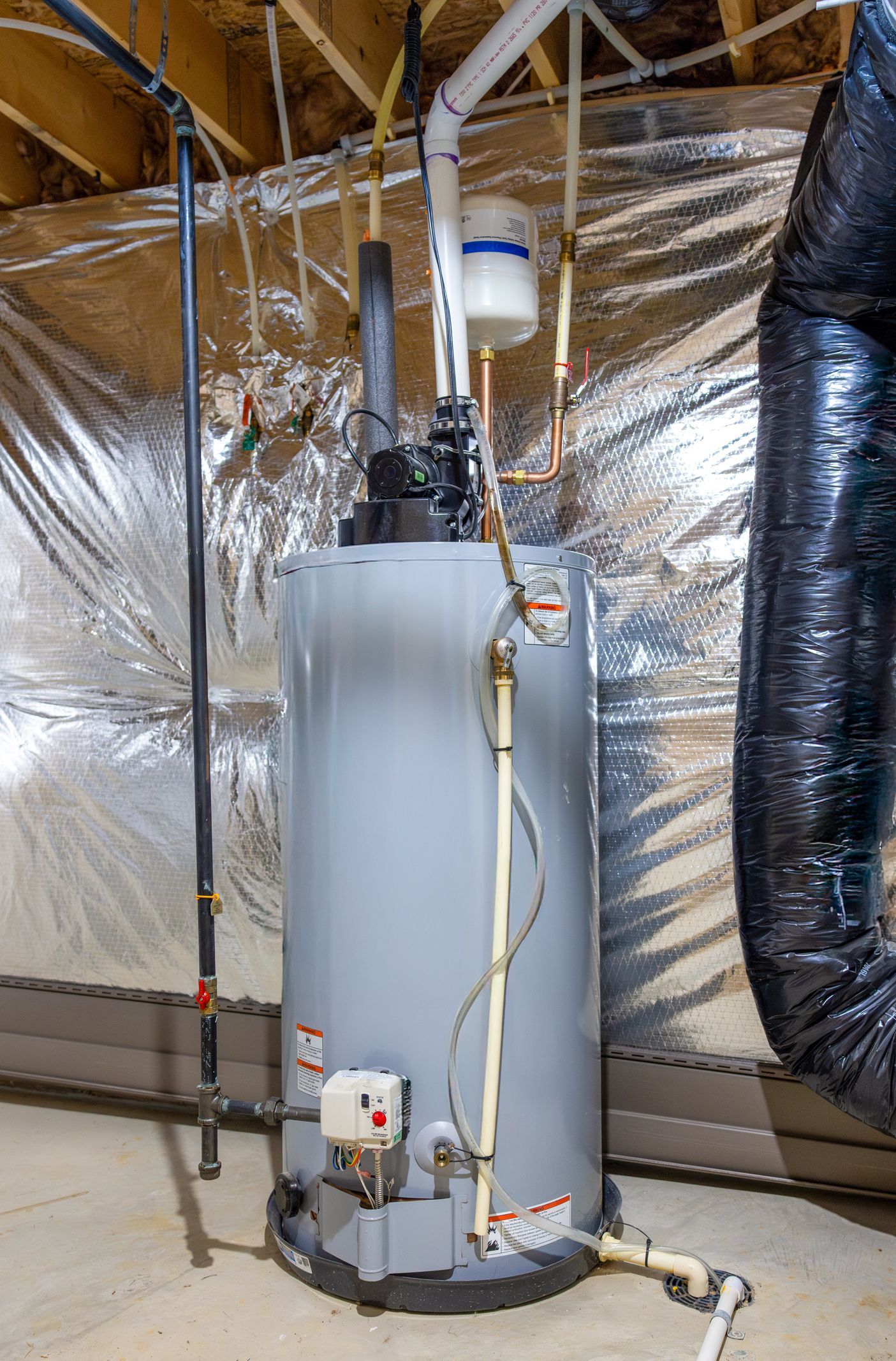 Tall white water heater with pipes in an unfinished attic, wrapped ducting beside it.