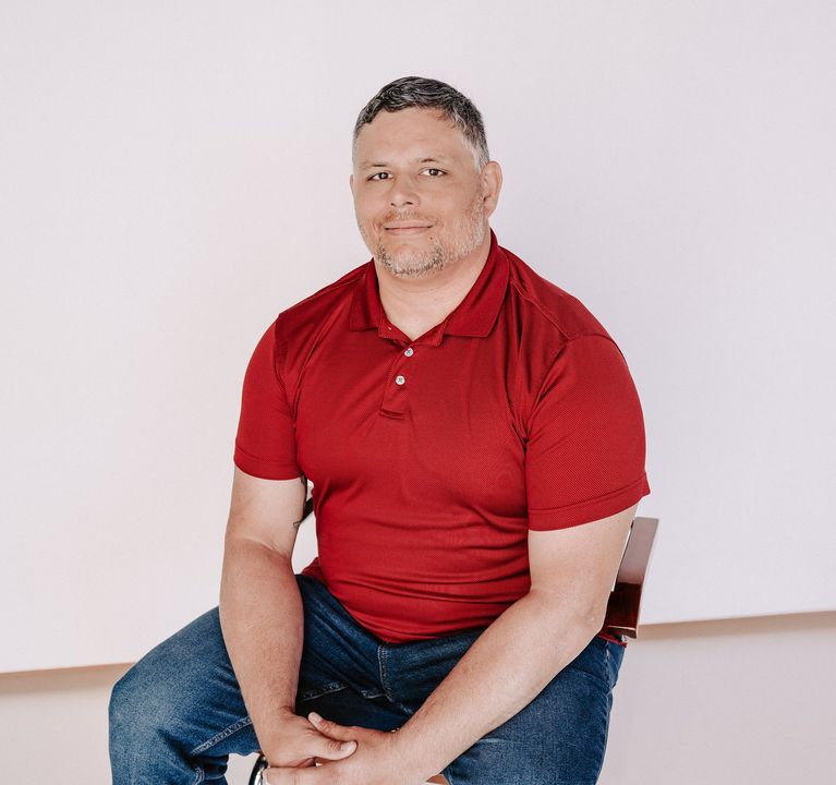 Man seated on a chair, wearing a red patterned shirt and jeans, against a plain light background