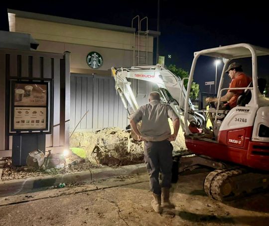 Night construction crew using a small excavator beside a building and fence, with a worker standing nearby.