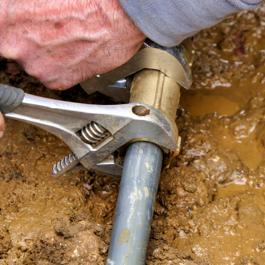 Close-up of a hand using an adjustable wrench to tighten a metal pipe fitting in muddy ground