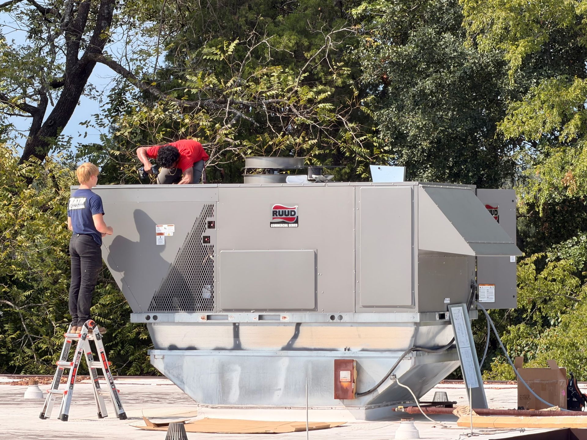 Workers on a large gray industrial unit outdoors, one on a ladder and one on top, with trees behind.