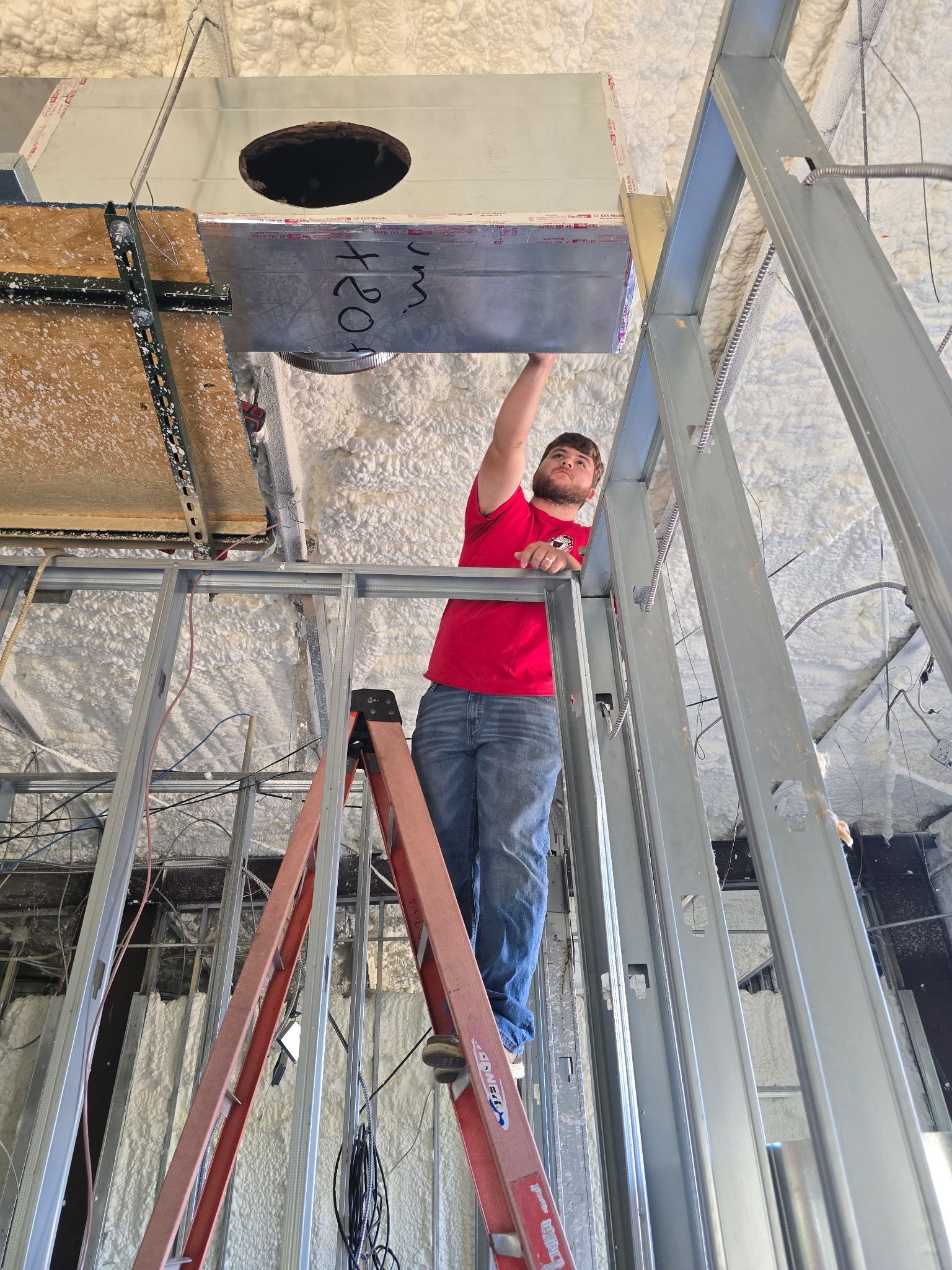 Worker in red shirt installing ceiling ducting inside a construction site, standing on ladders.