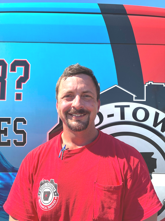 Man smiling in a red T-shirt standing in front of a blue and red vehicle with a logo.