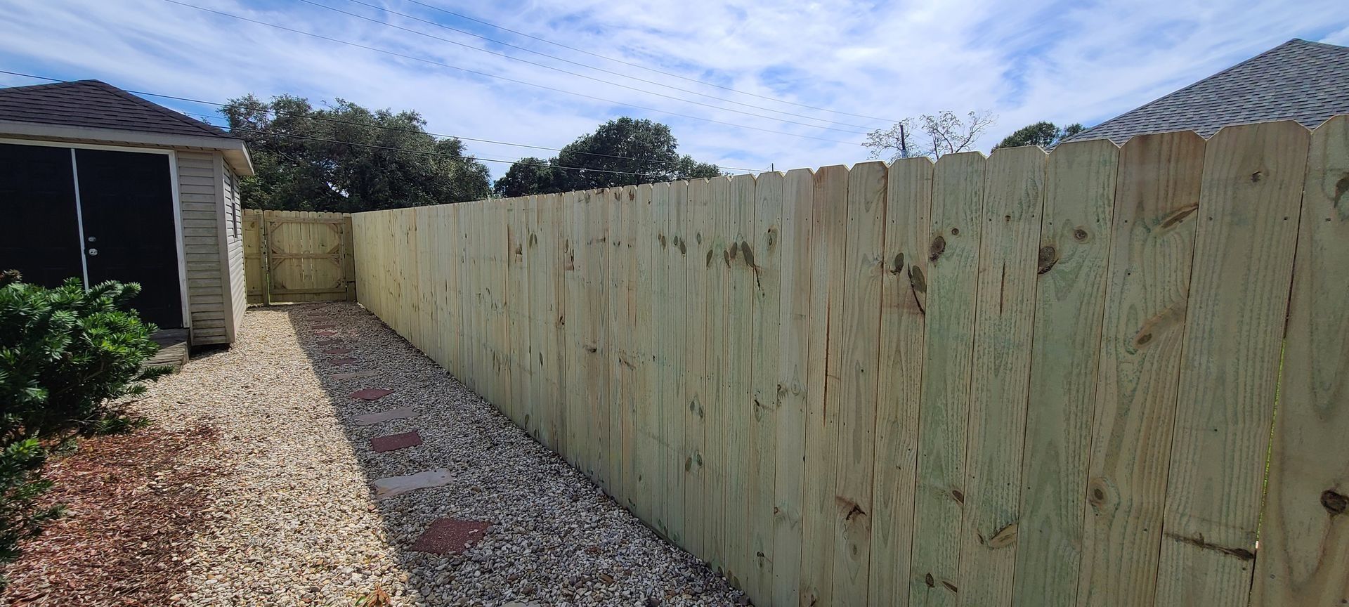 Long wooden fence along a gravel side yard beside a house under a blue sky