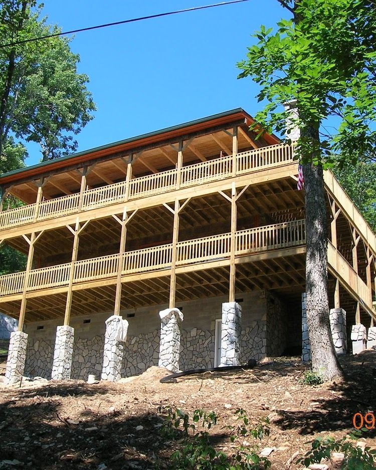 Three-story wooden lodge on a stone foundation amid trees on a sunny hillside