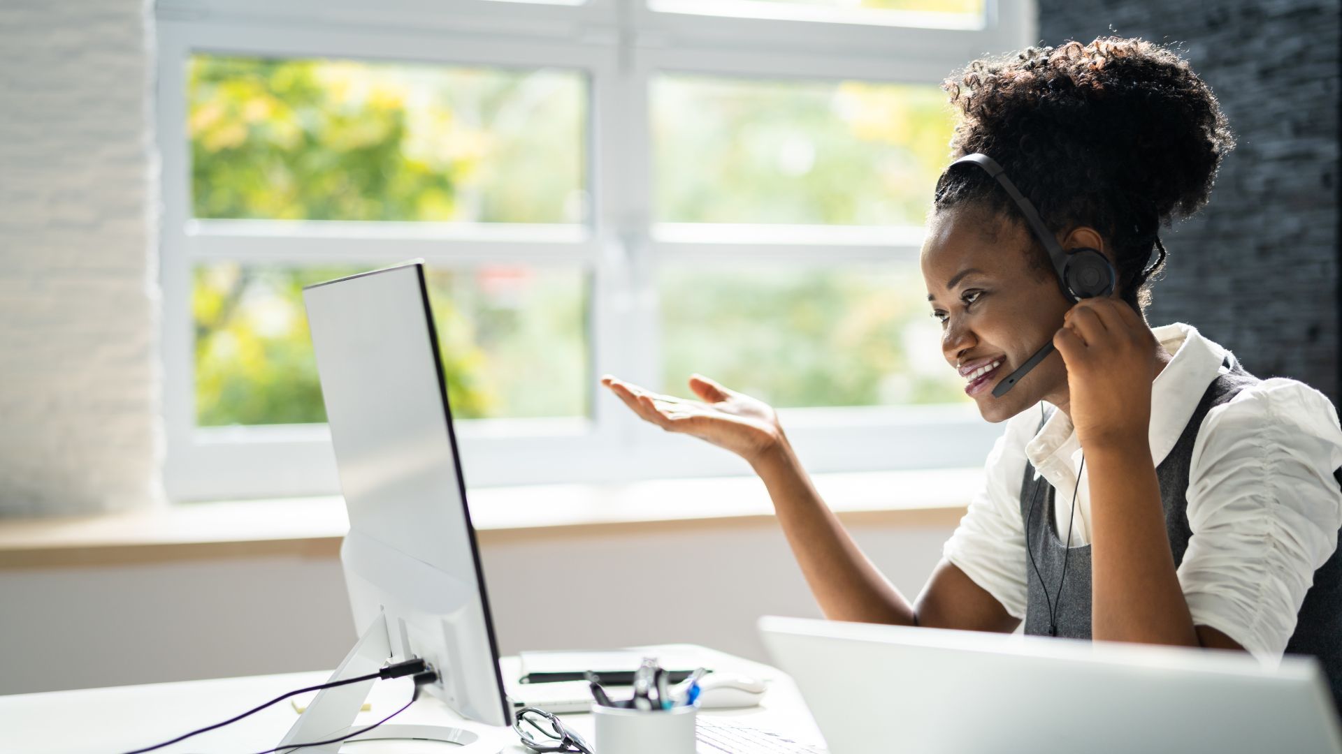 A woman wearing a headset gestures while looking at a computer screen, near a window.