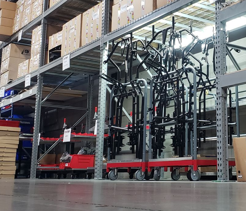 Warehouse aisle with red carts, stacked boxes on metal shelving, and folded black chairs under bright lights