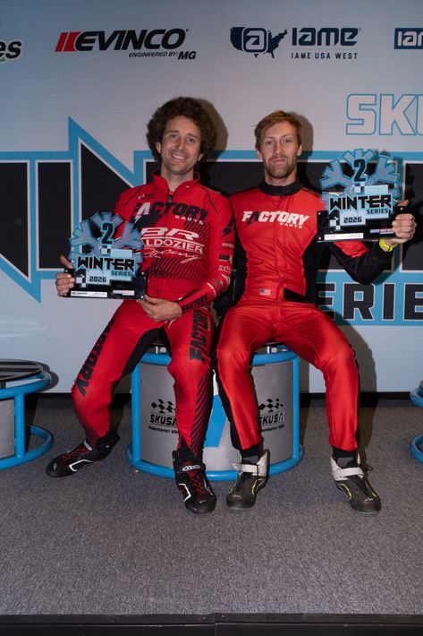 Two racers in red suits posing on a podium with trophies against a motorsport backdrop