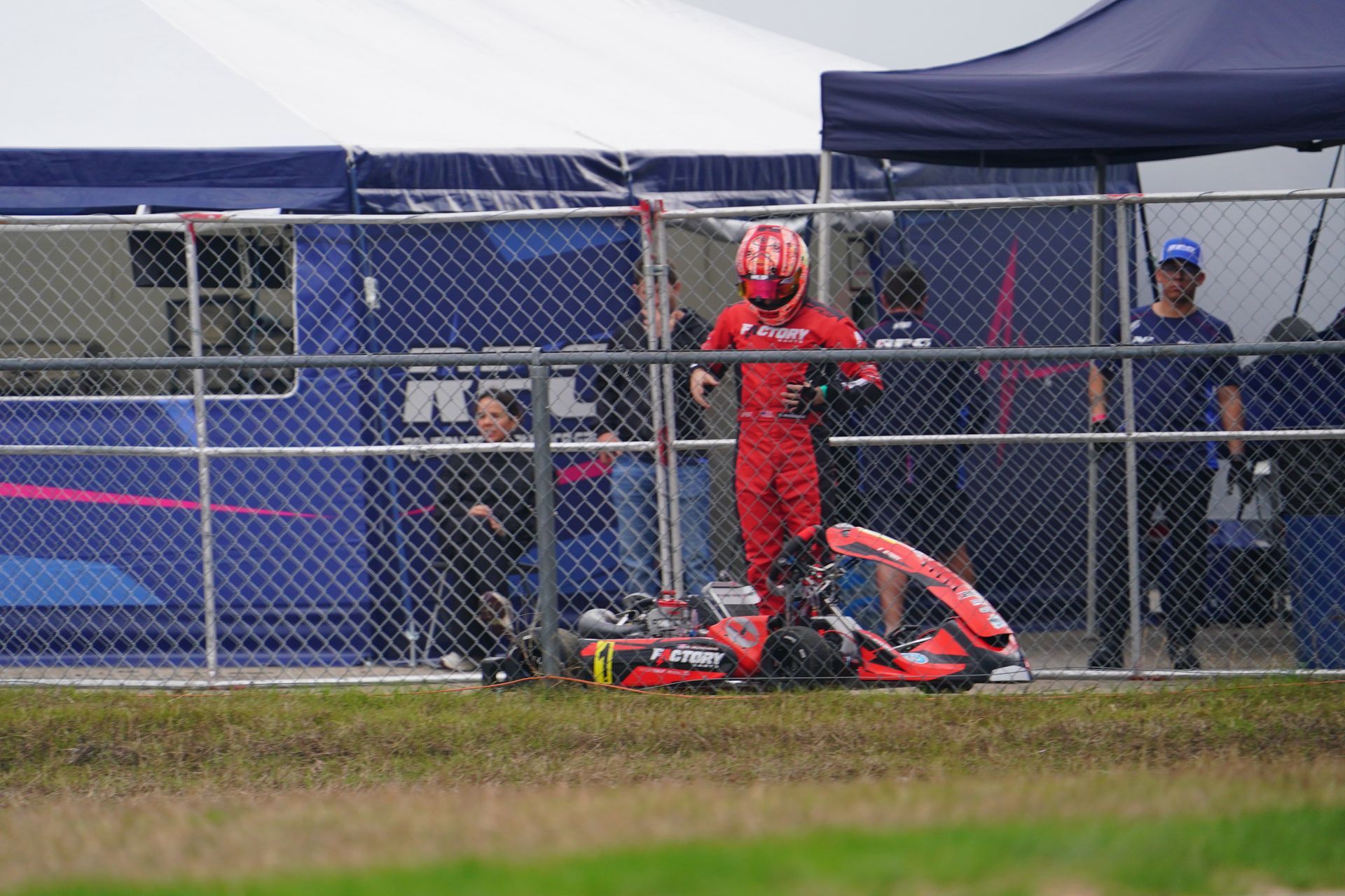Racer in red suit standing beside a crashed red go-kart near a chain-link fence and tent