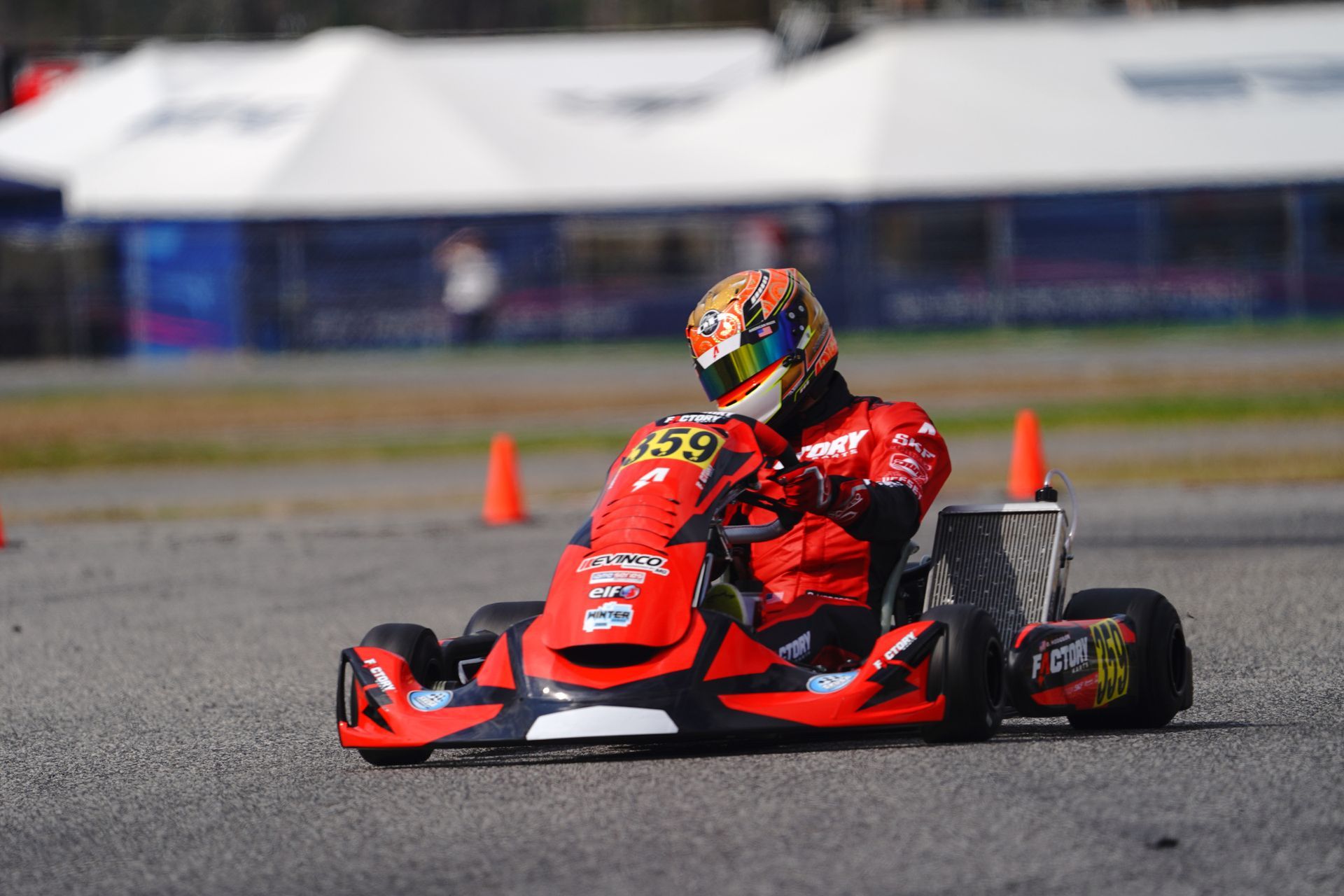Red go-kart racer speeding on a track, wearing a helmet and red suit, with orange cones nearby