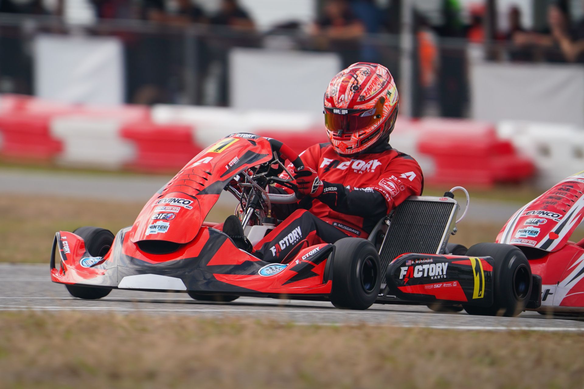 Red go-kart racer speeding on a wet track, wearing red gear and helmet, with blurred barriers behind.