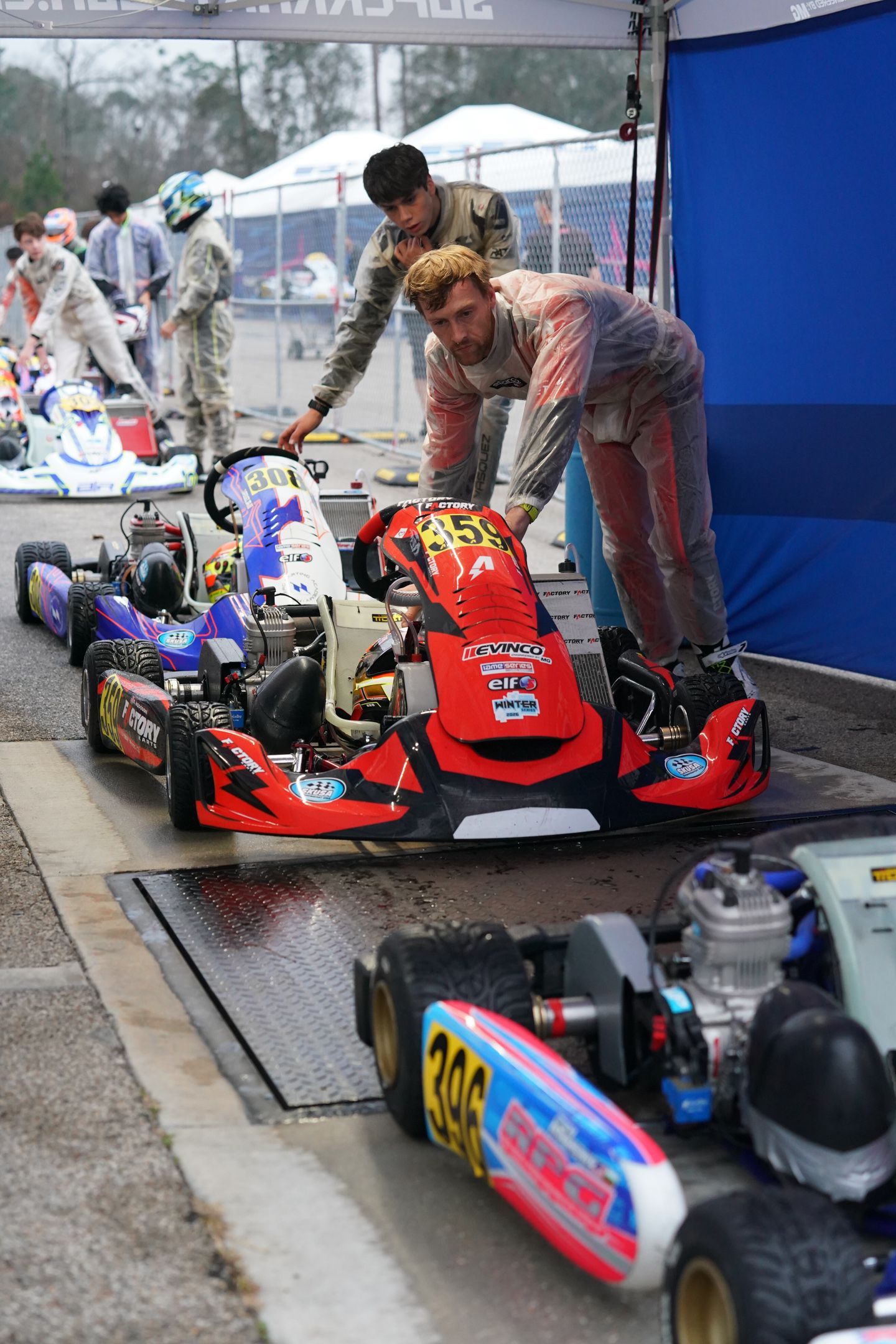 Go-karts lined up in pit area, with people adjusting a red kart under a blue tent.