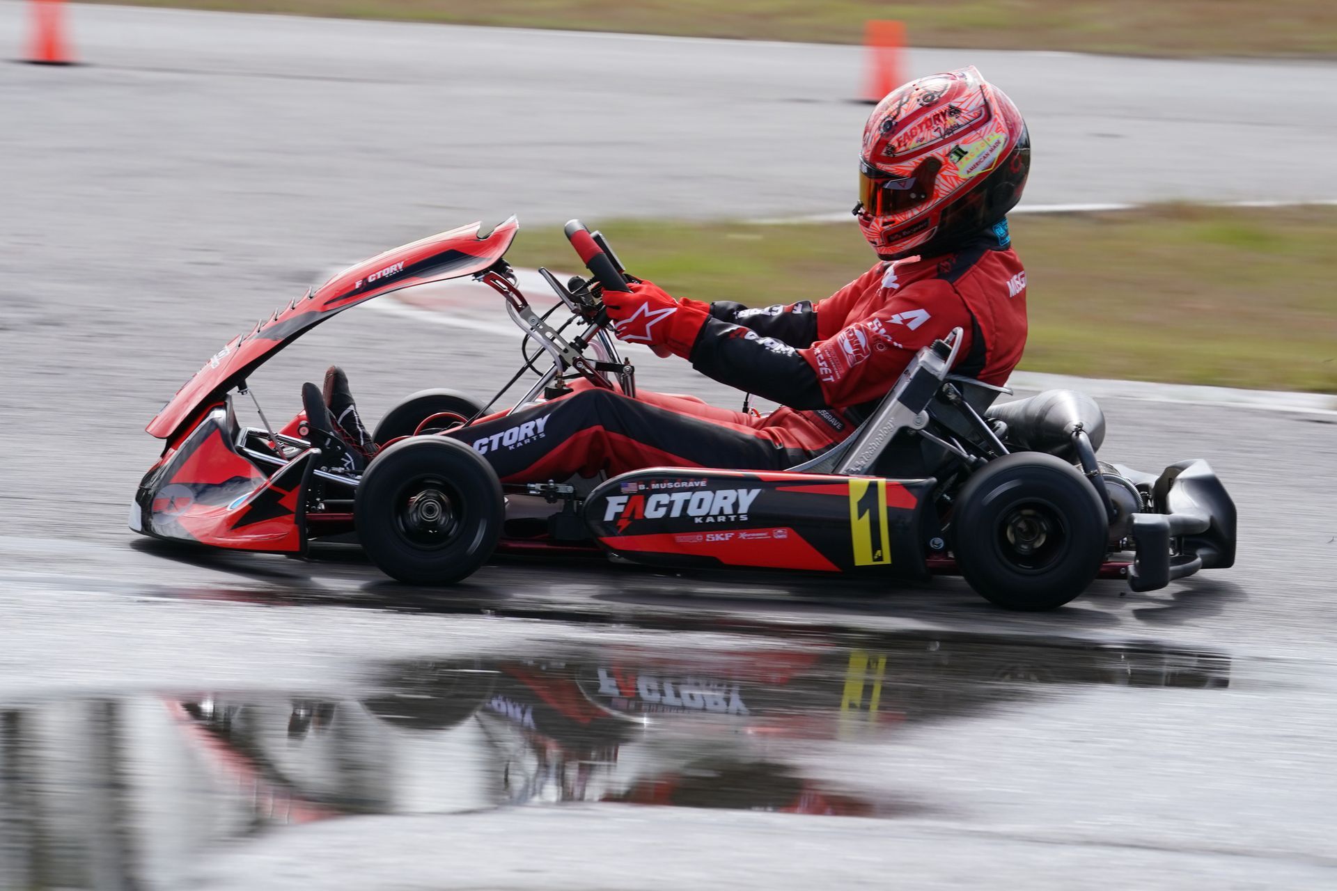 Red go-kart racer speeding on a wet track, wearing red gear and helmet, with spray from the tires