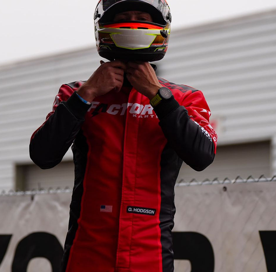 Race driver in a red suit fastening a black helmet on a track backdrop