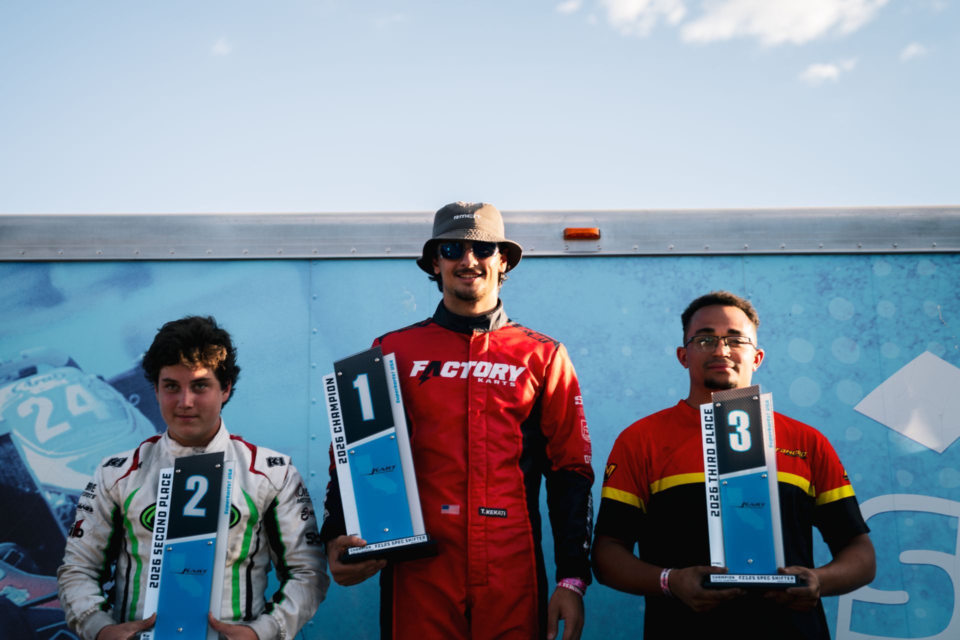 Three shifter kart racers on a podium holding trophies, with Theo Kekati in red on top P1
