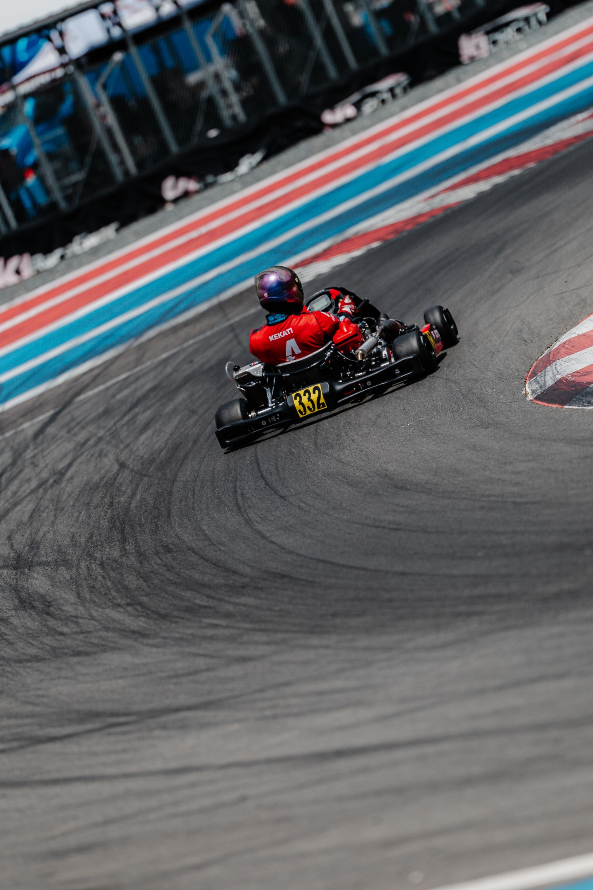 Racer in red suit standing beside a crashed red go-kart near a chain-link fence and tent