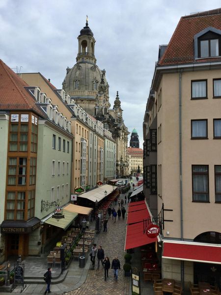 Narrow European street with pedestrians, colorful buildings, and a tall ornate church tower in the distance