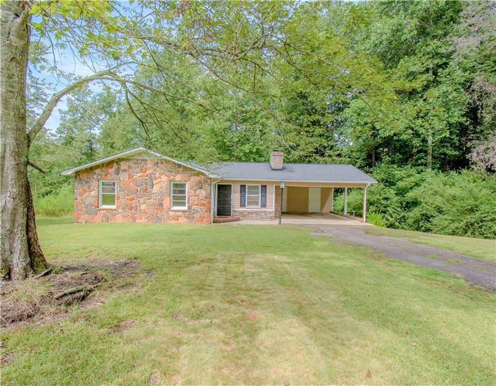 Single-story brick house with a carport, set on a grassy lot surrounded by trees.
