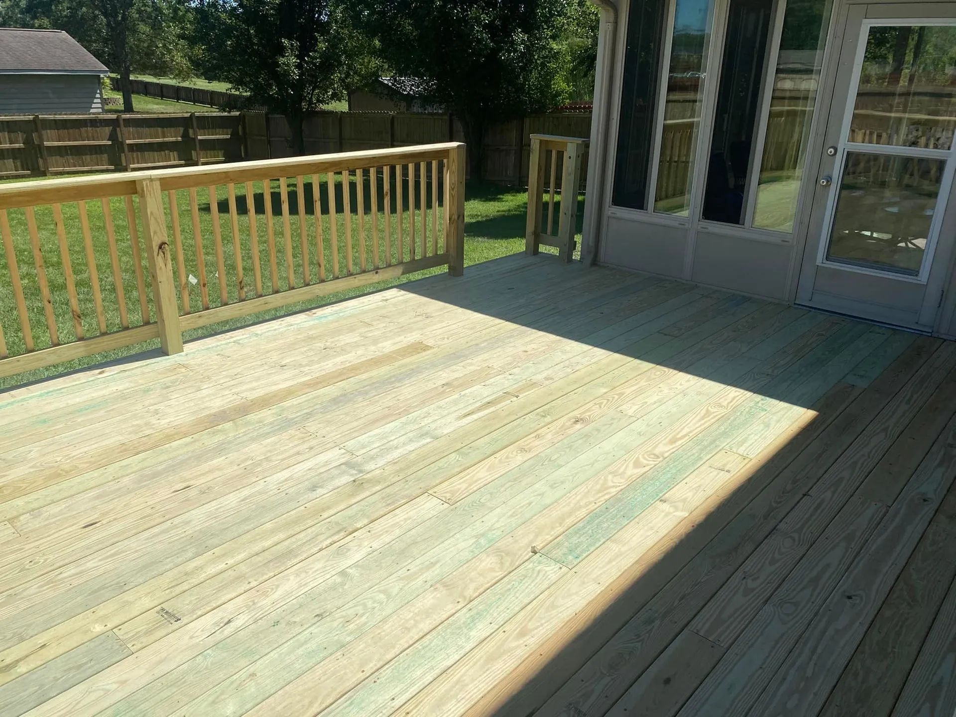Sunny wooden deck with railing beside a house, overlooking a grassy yard.