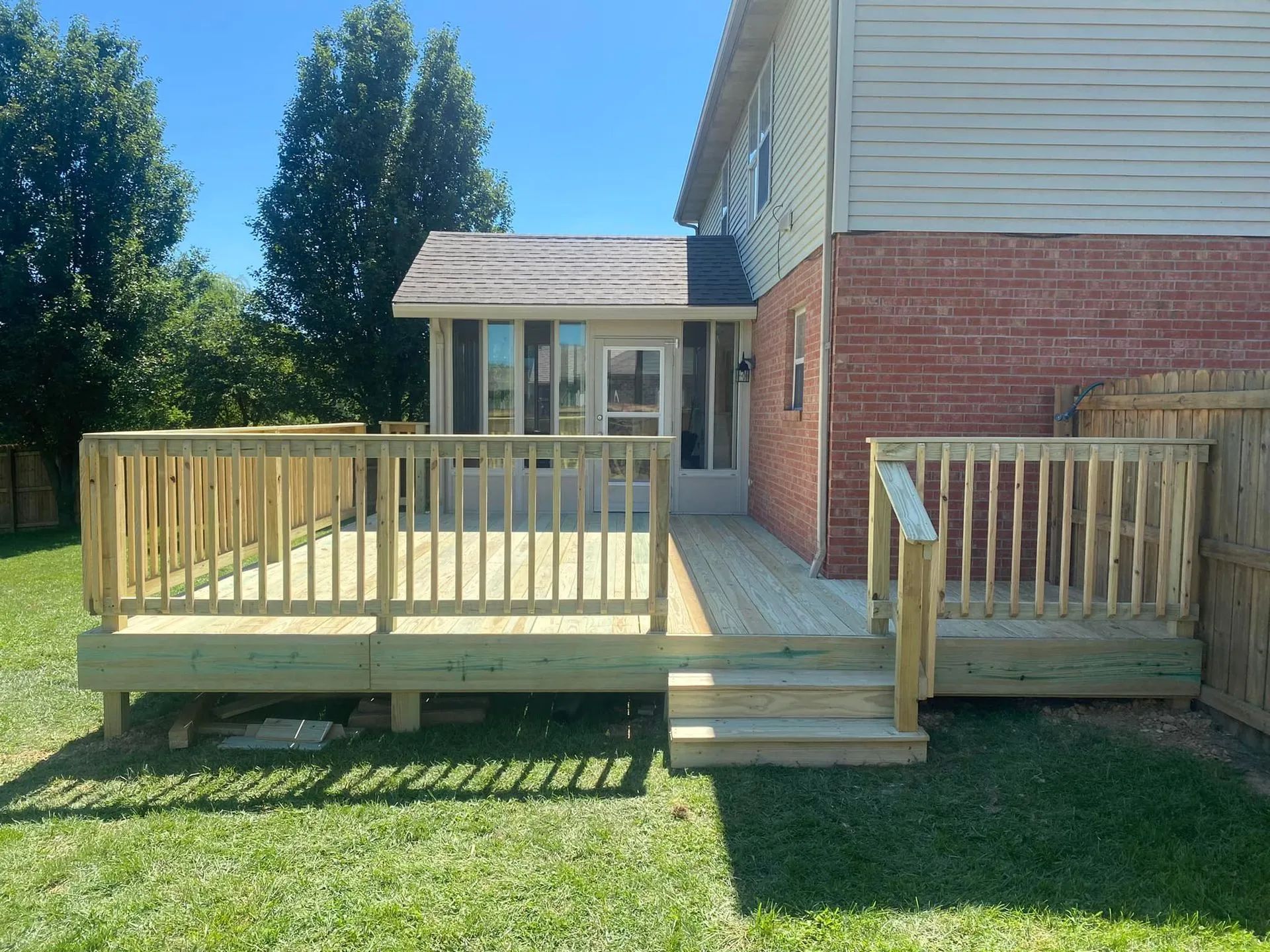 A newly built wooden deck with railings and steps attached to the rear of a brick house with a sunroom.