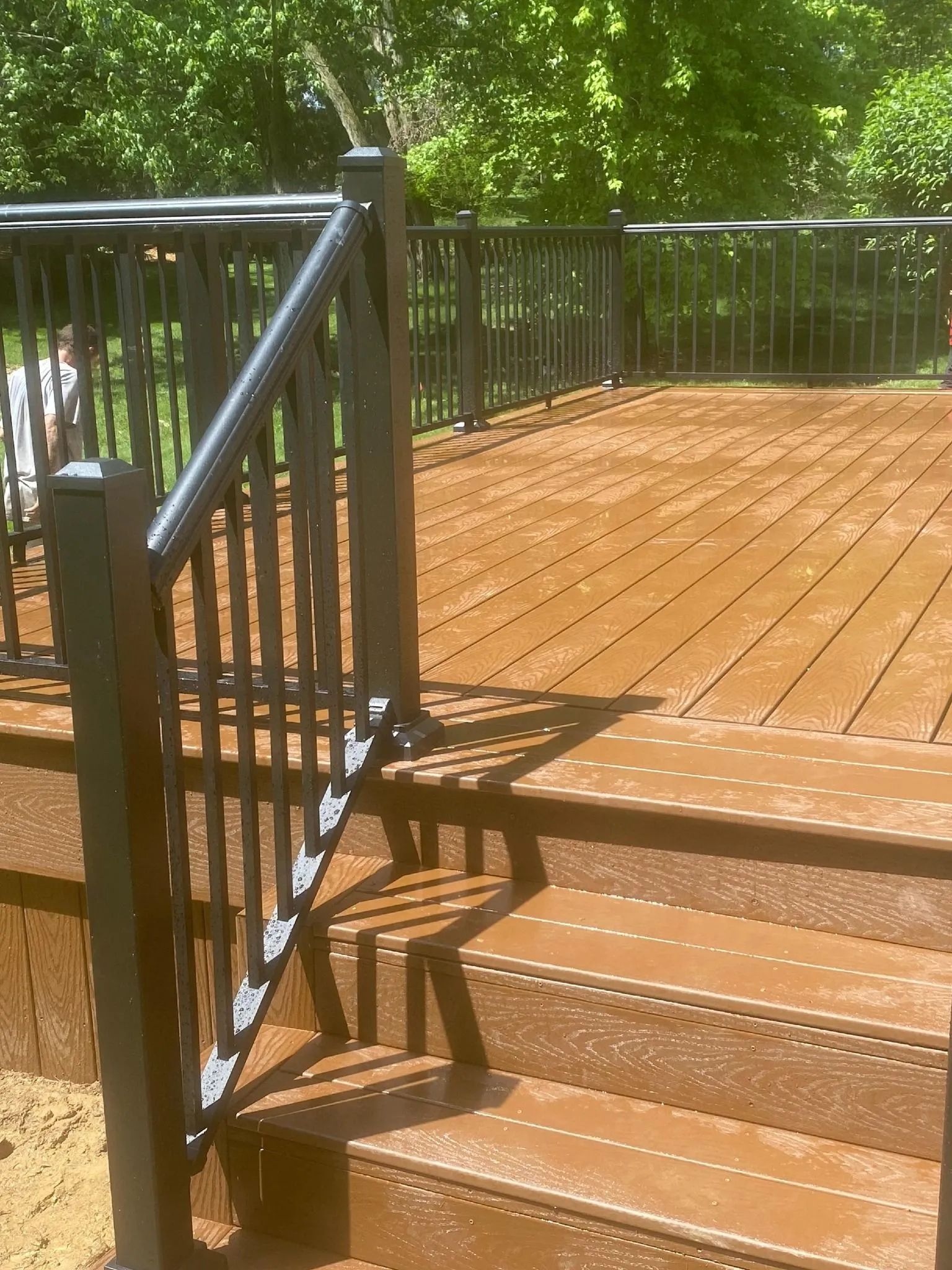 Wooden deck with black metal railing and steps, surrounded by green trees and sunlight