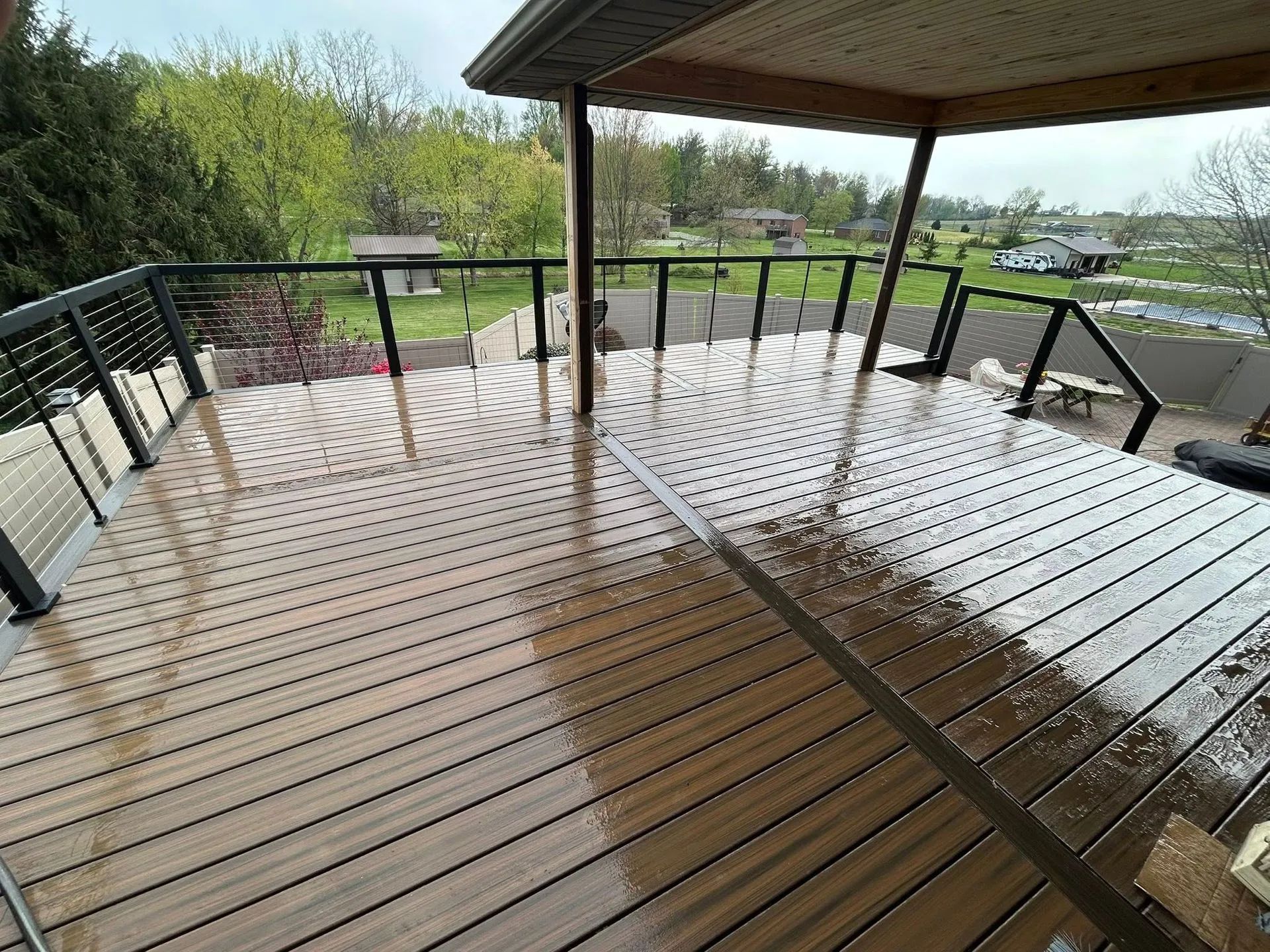 Wet wooden deck with black railing overlooking a green yard under a roofed patio.