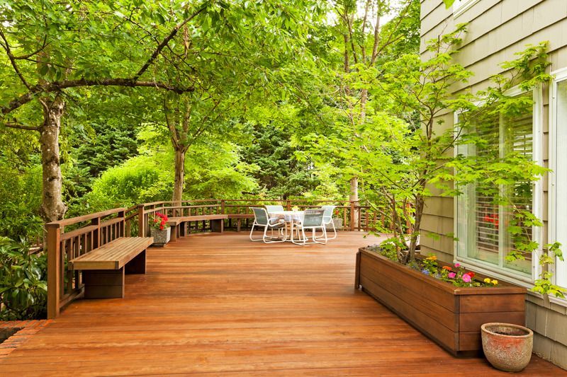 Wooden deck with trees, benches, white table set, and a house wall with windows and planters