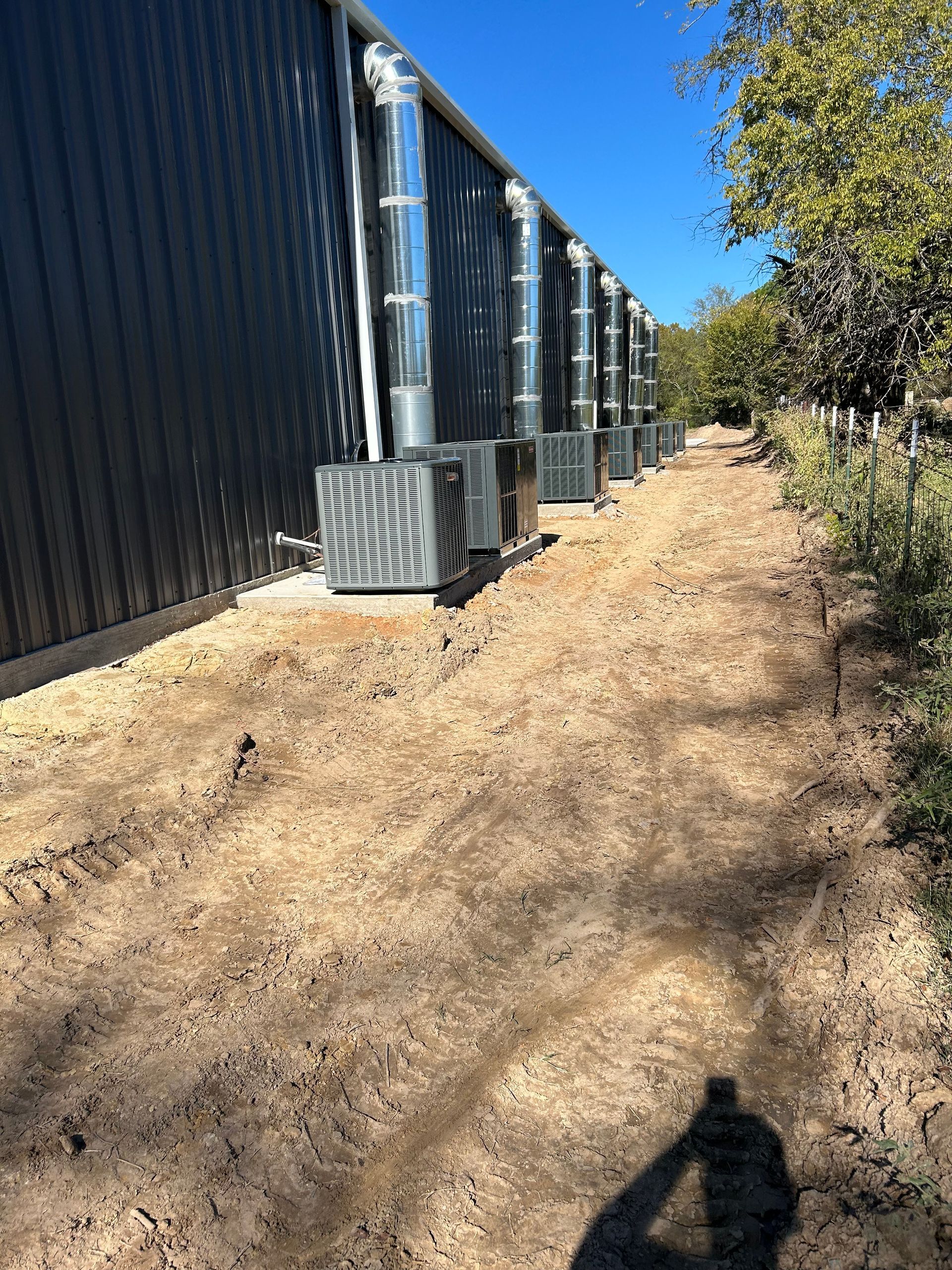 Air conditioning units line the side of a dark metal building, with silver exhaust pipes. Sunny, dirt path.