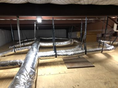 HVAC ductwork in an attic, suspended from beams. Aluminum flexible tubing, plywood floor, dark rafters.