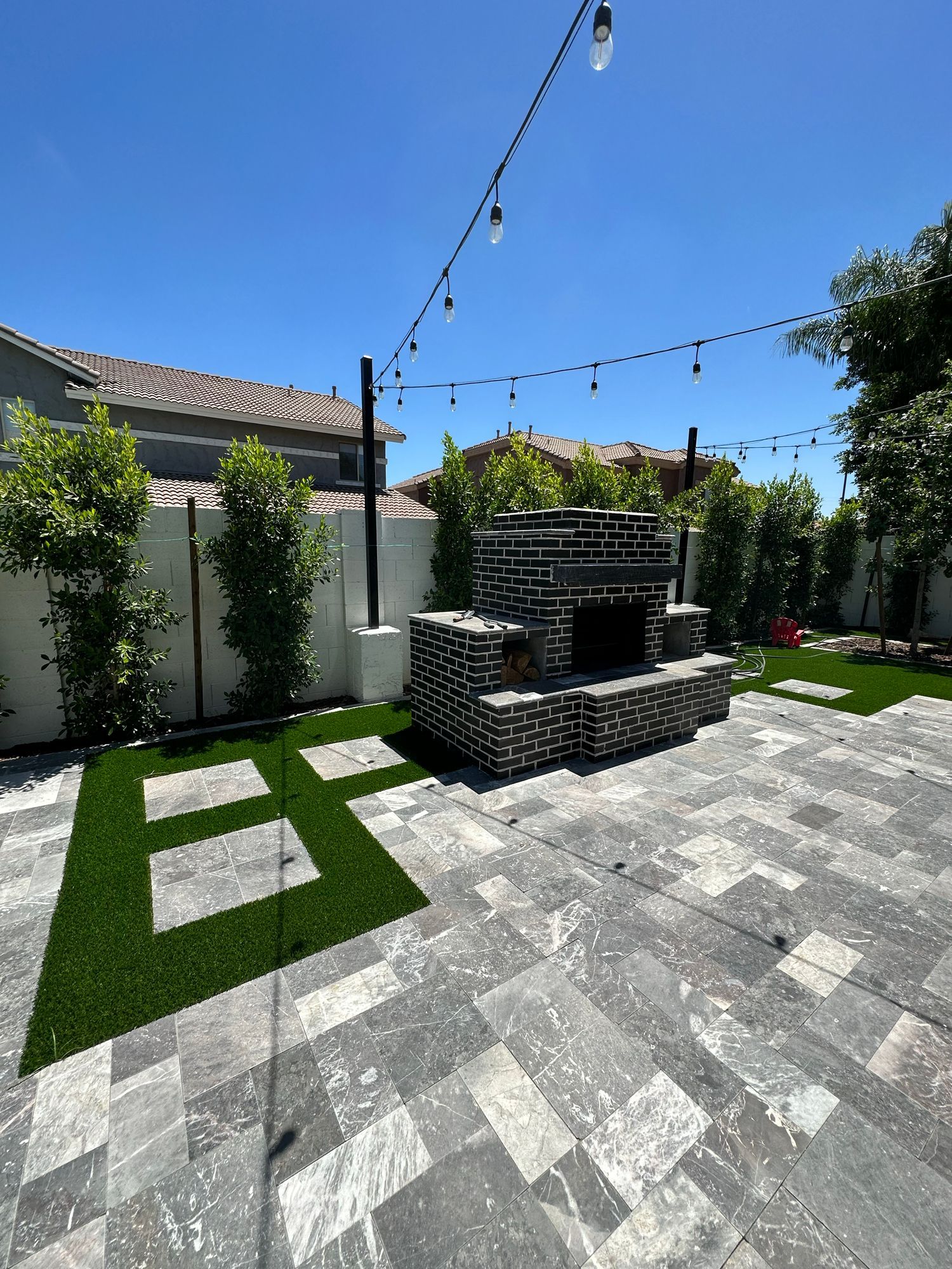 A brick outdoor fireplace on a grey stone patio with grass accents, framed by tall green shrubs under string lights.