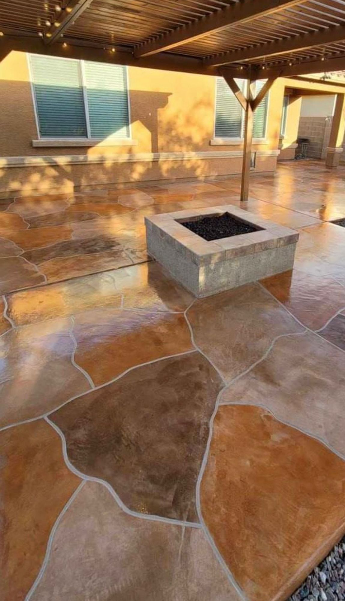 A patio with multicolored, irregular stone-look concrete tiles featuring a square fire pit under a wooden pergola.