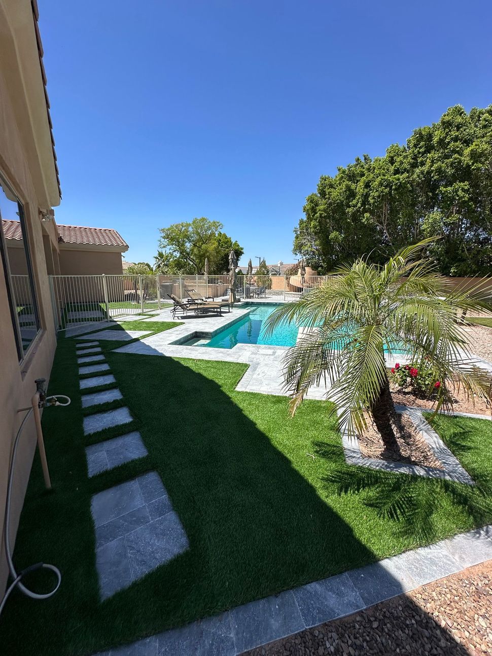 A sunny backyard featuring a rectangular swimming pool, stepping stones through a lawn, and mature trees under a blue sky.