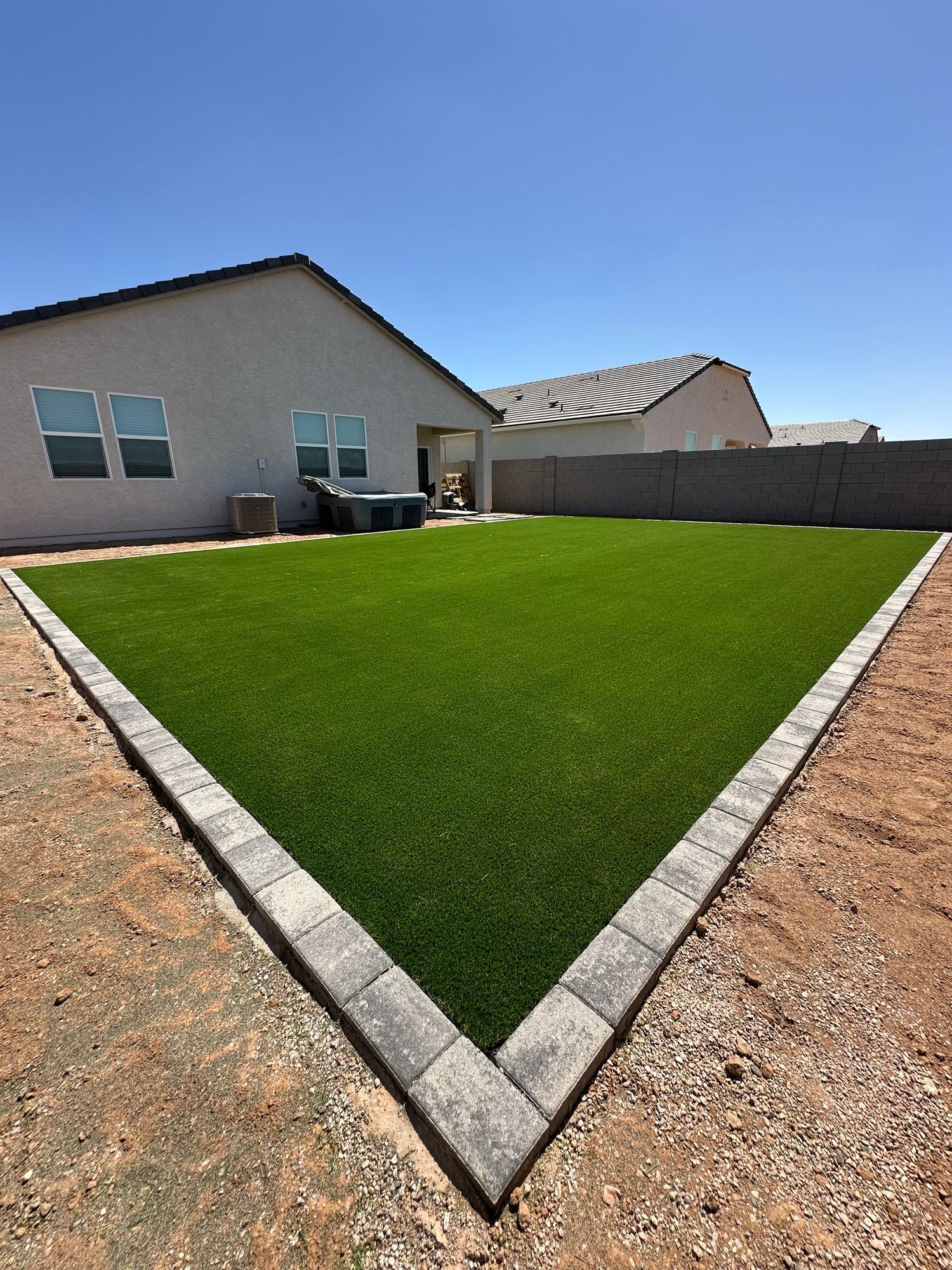 A rectangular patch of green artificial grass bordered by grey pavers in a backyard with beige houses and gravel.