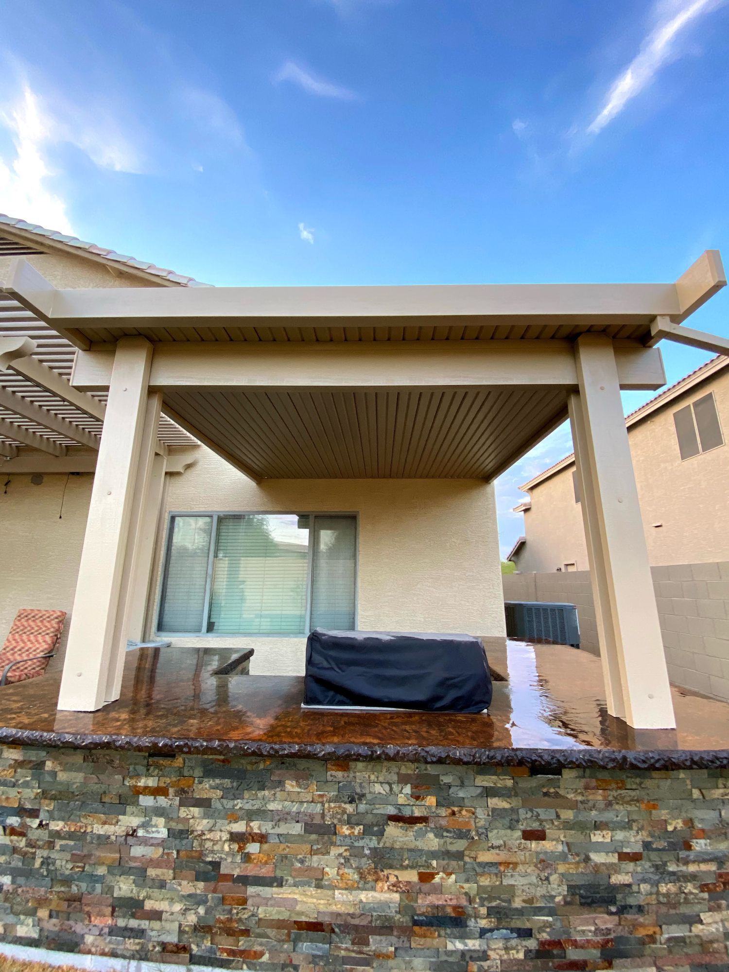 A stone-faced outdoor kitchen counter sits beneath a beige patio cover attached to a house under a blue sky.