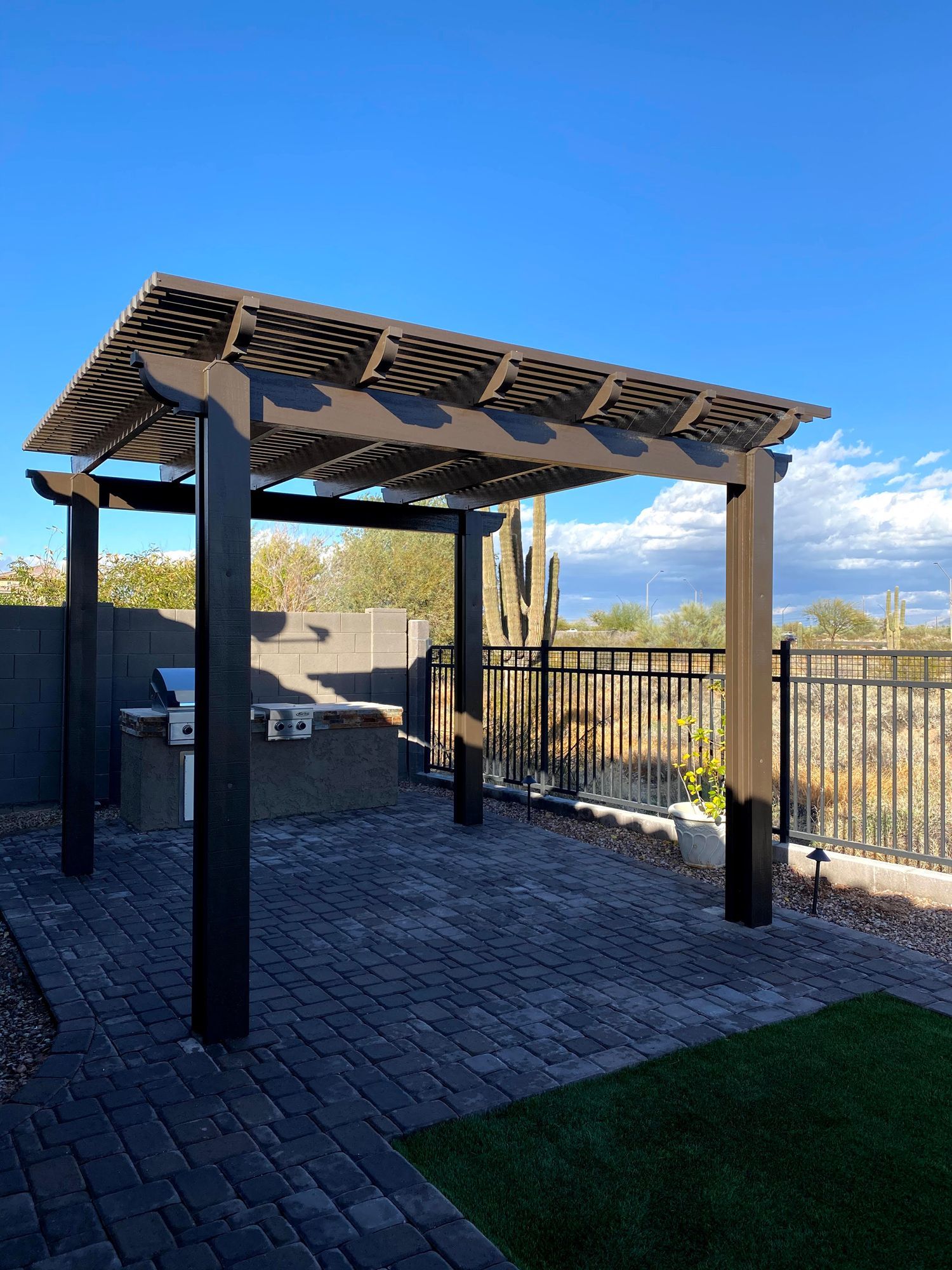 A black outdoor pergola stands over a brick patio with a built-in stone grill against a fence and desert background.