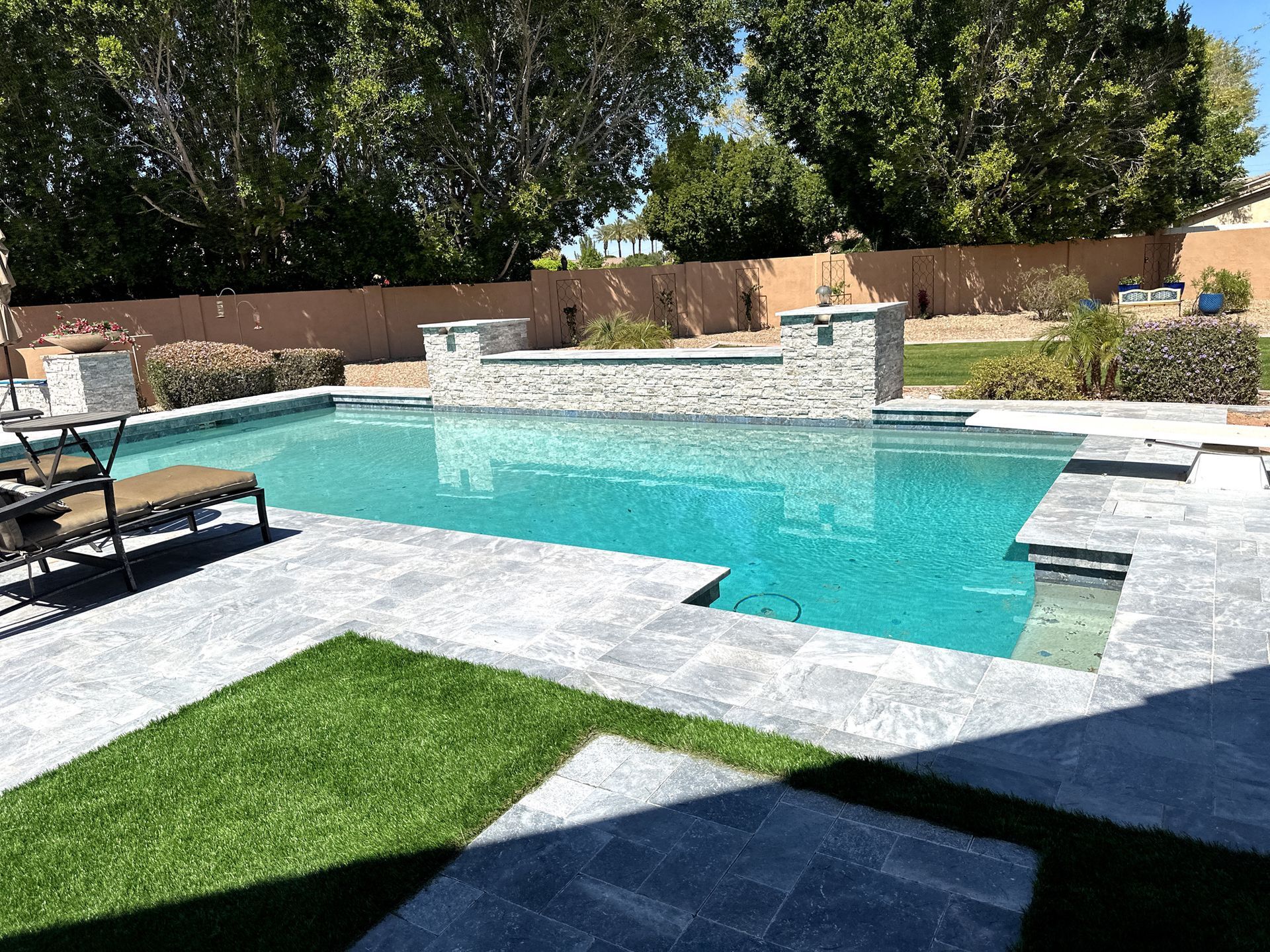 A bright blue swimming pool with a stone water feature, surrounded by grey pavers and a small lawn in a backyard.