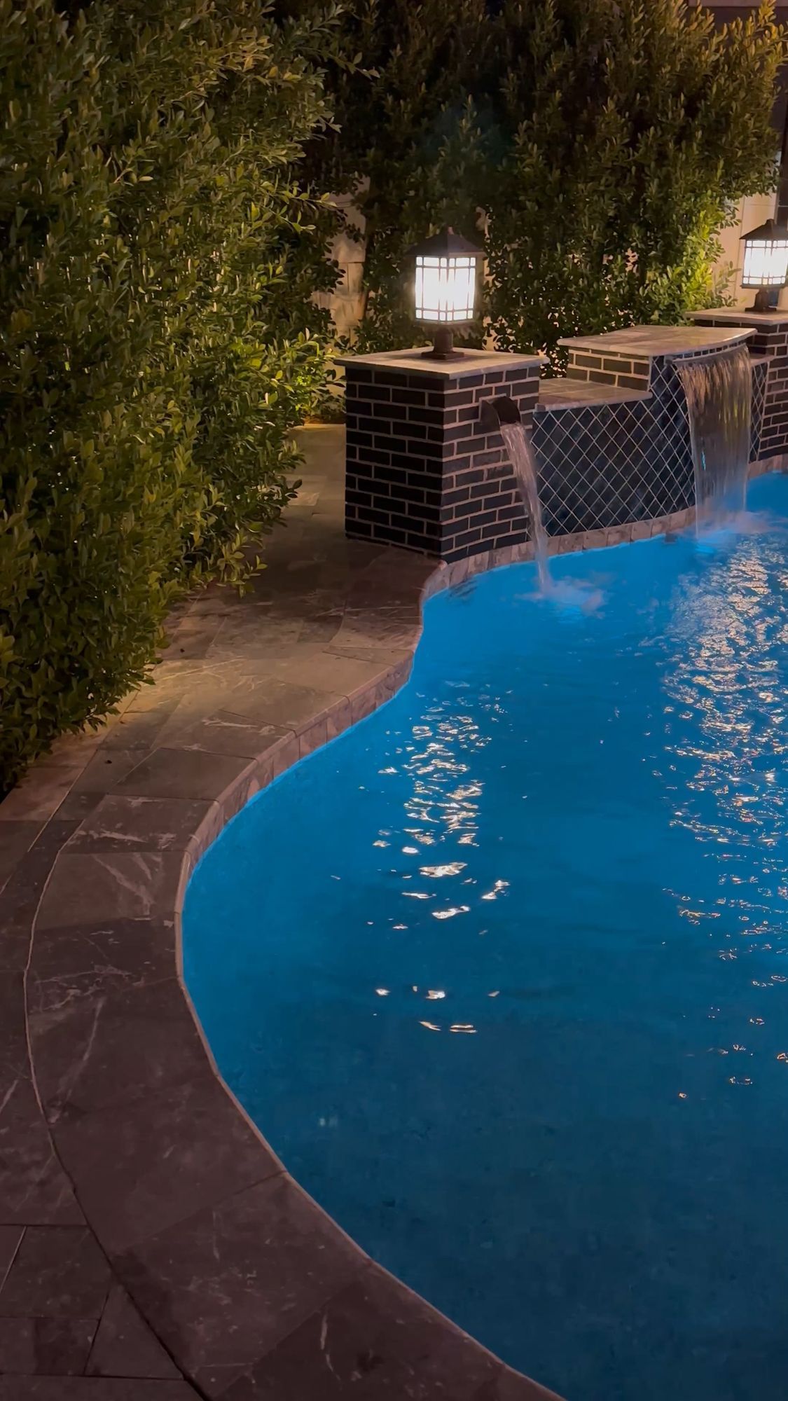 A blue swimming pool at night with water features flowing from stone pillars, surrounded by illuminated green hedges.