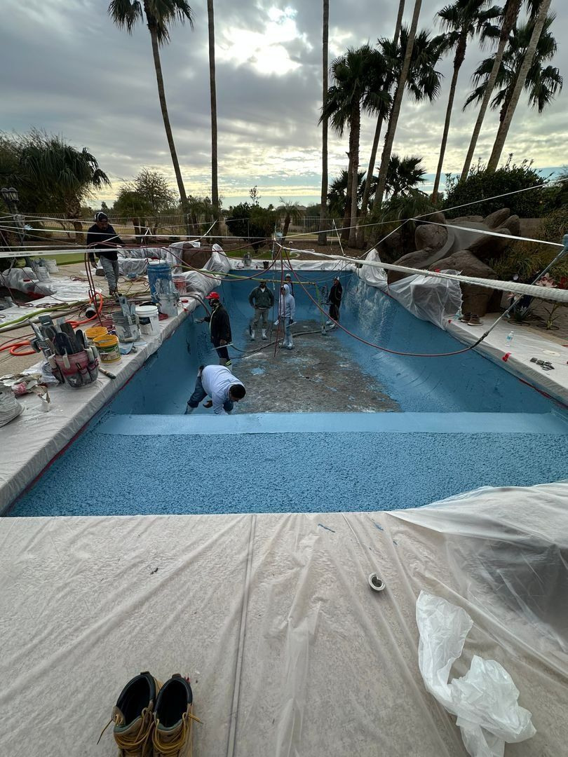 Workers renovate a backyard swimming pool, installing blue tiles while surrounded by construction tools and materials.